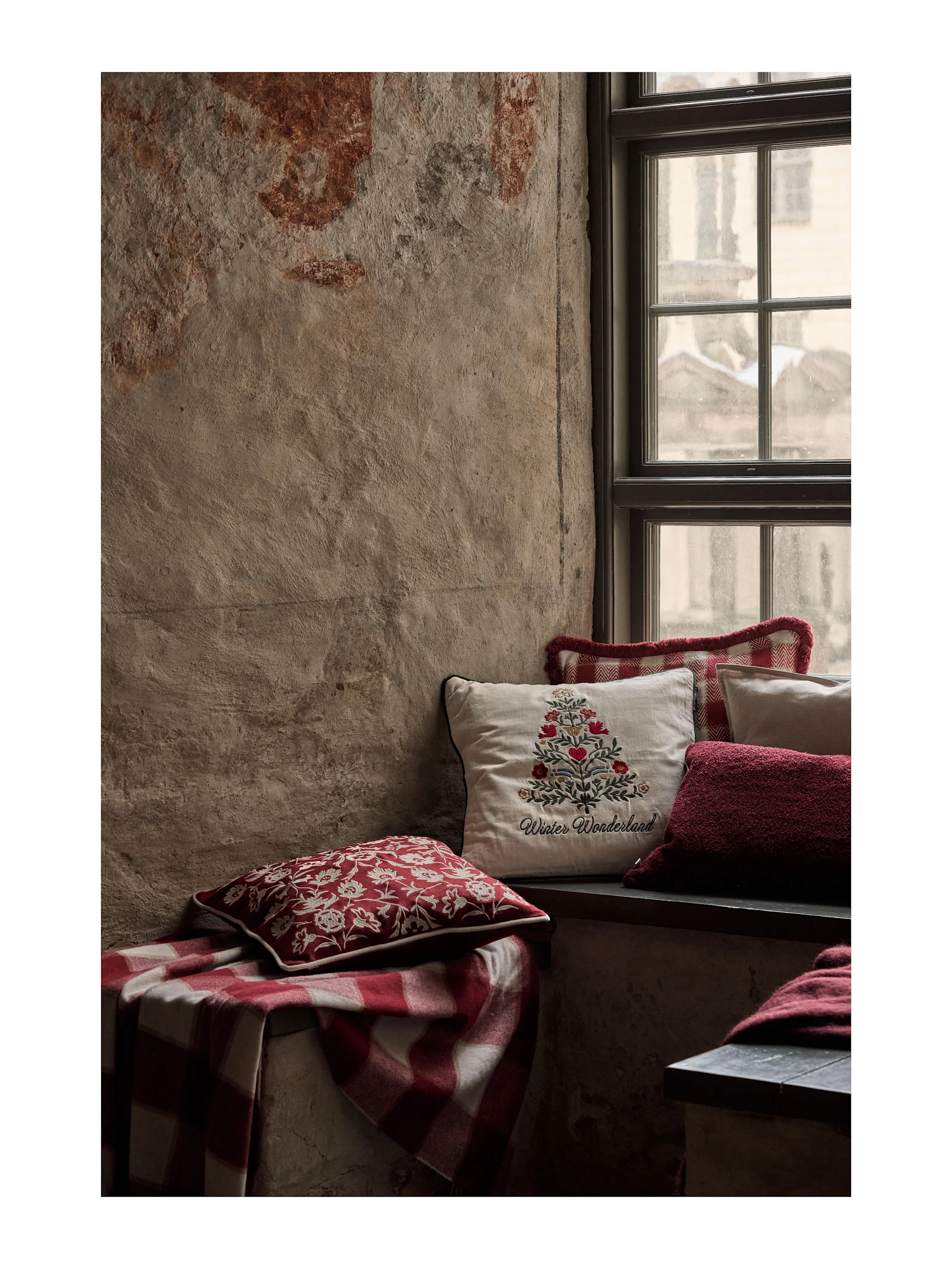 Gemütlicher Fenstersitz mit festlichen rot-weißen Kissen und einer karierten Decke. Ein cremefarbenes Kissen zeigt 'Winter Wonderland' Stickerei.