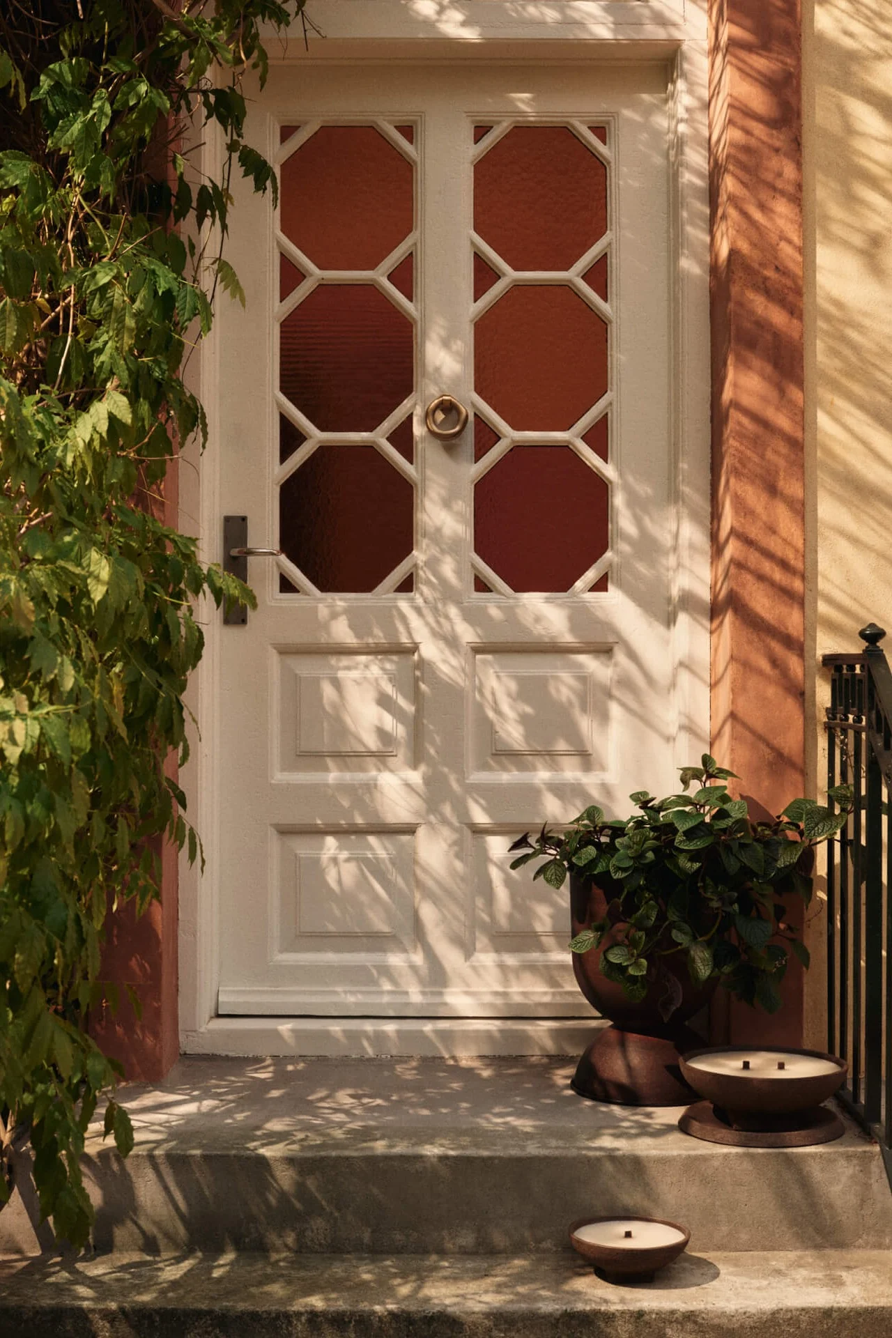 Weiße Haustür mit achteckigen Glaspaneelen, grünen Ranken links und einem Pflanzgefäß mit zwei Kerzen auf den Stufen.