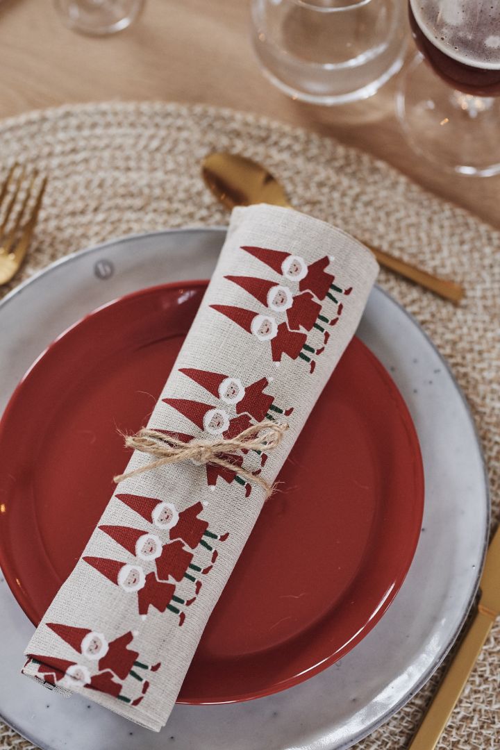 Ein zusammengerolltes Geschirrtuch mit Weihnachtsmann-Motiven ruht auf einem roten Teller und einem Nordic Sand Teller von Broste Copenhagen.