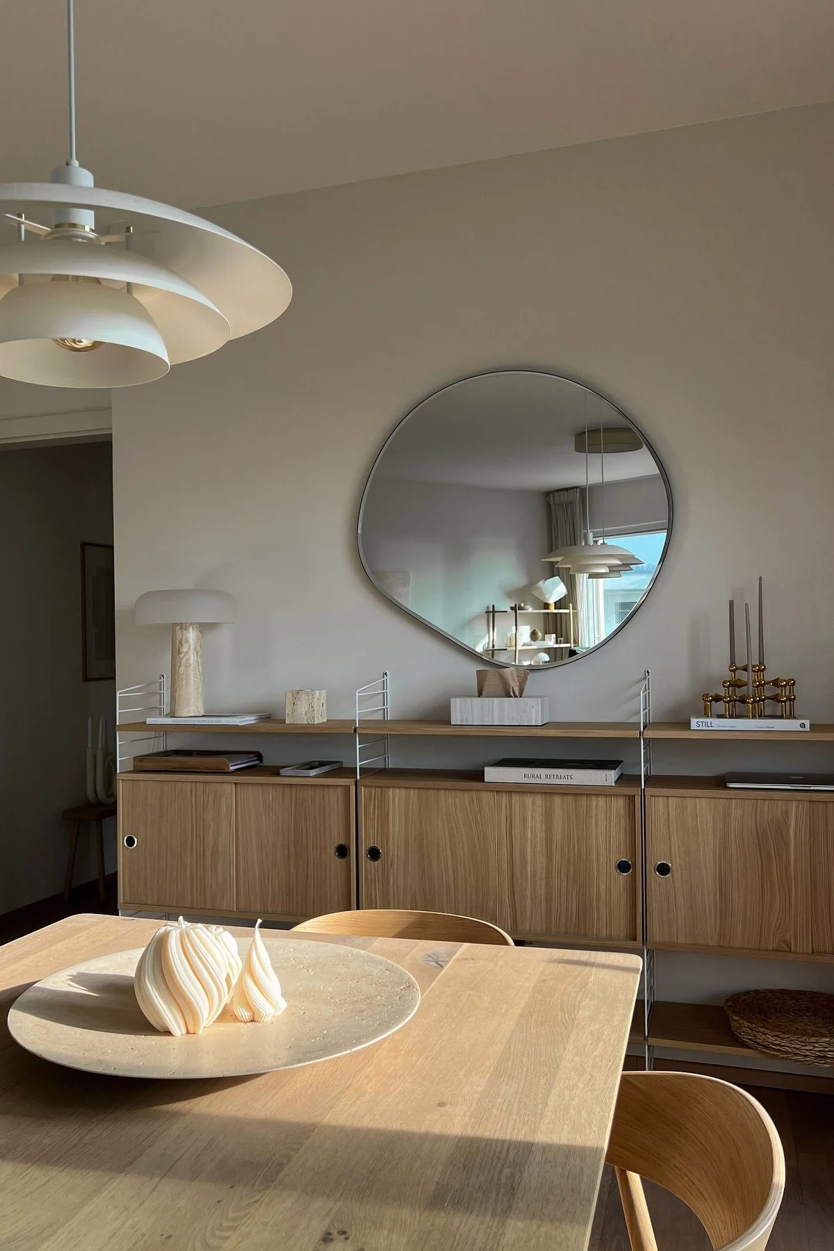 Modernes Esszimmer mit Holztisch, zwei Spiralkerzen, Holzregal, unregelmäßigem Spiegel und gestufter Lampe.