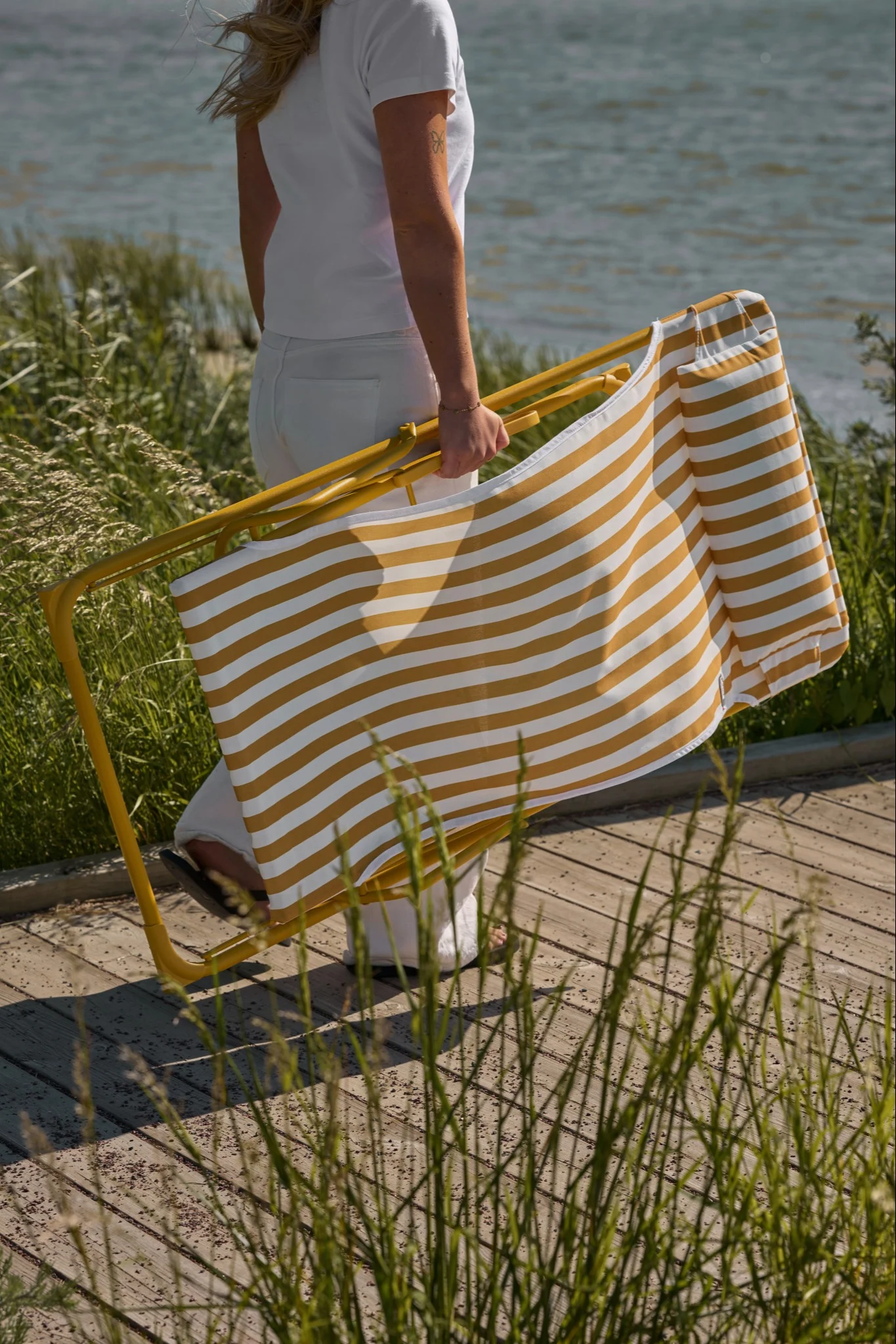 Person in Weiß trägt einen zusammengeklappten gelb-weiß gestreiften Strandstuhl auf einem Holzsteg am Wasser mit hohem Gras.