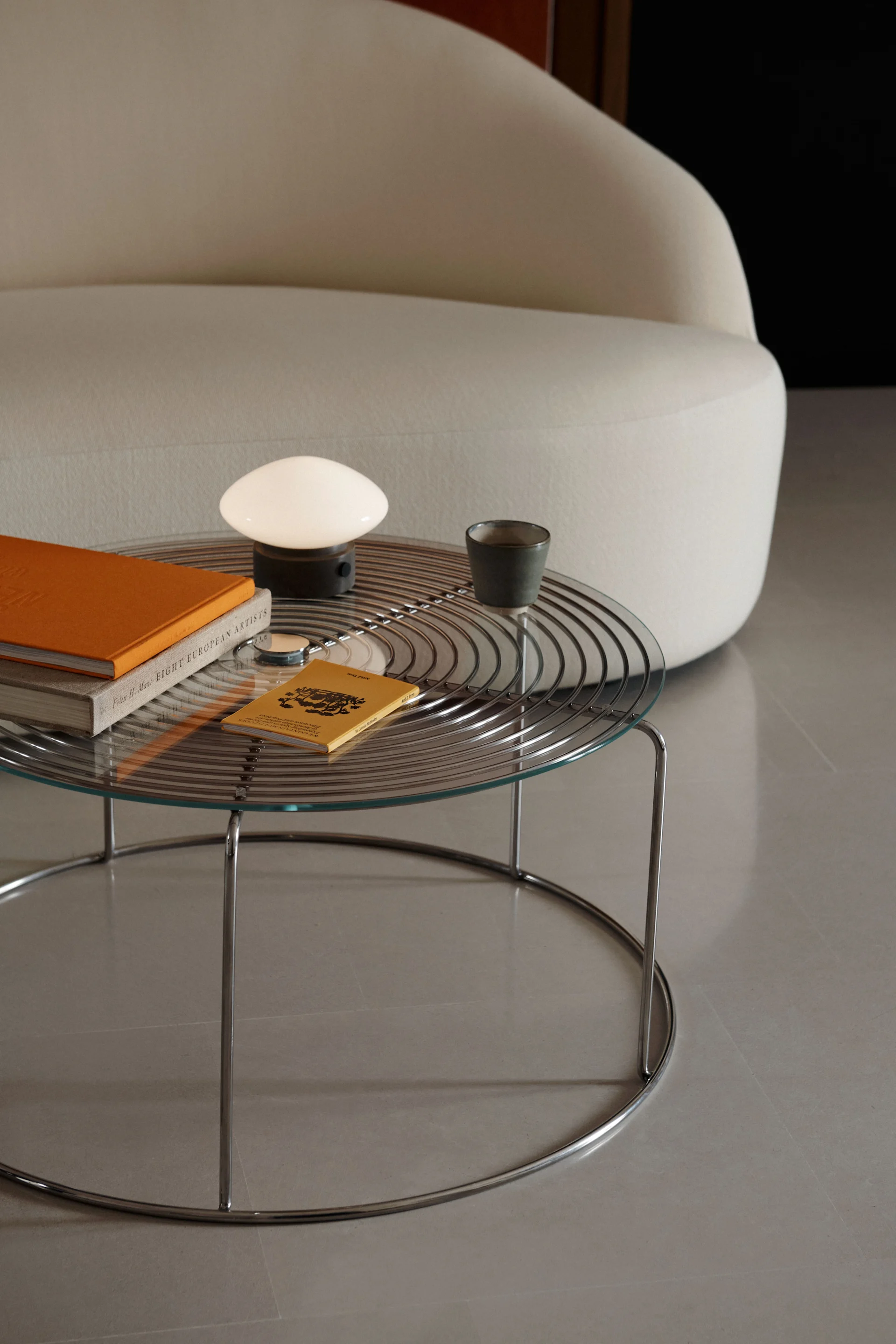 Ein Couchtisch mit Glasplatte und Chromgestell, auf dem eine weiße Pilzlampe, ein orangefarbenes Buch und eine dunkle Tasse neben einem geschwungenen cremefarbenen Sofa stehen.