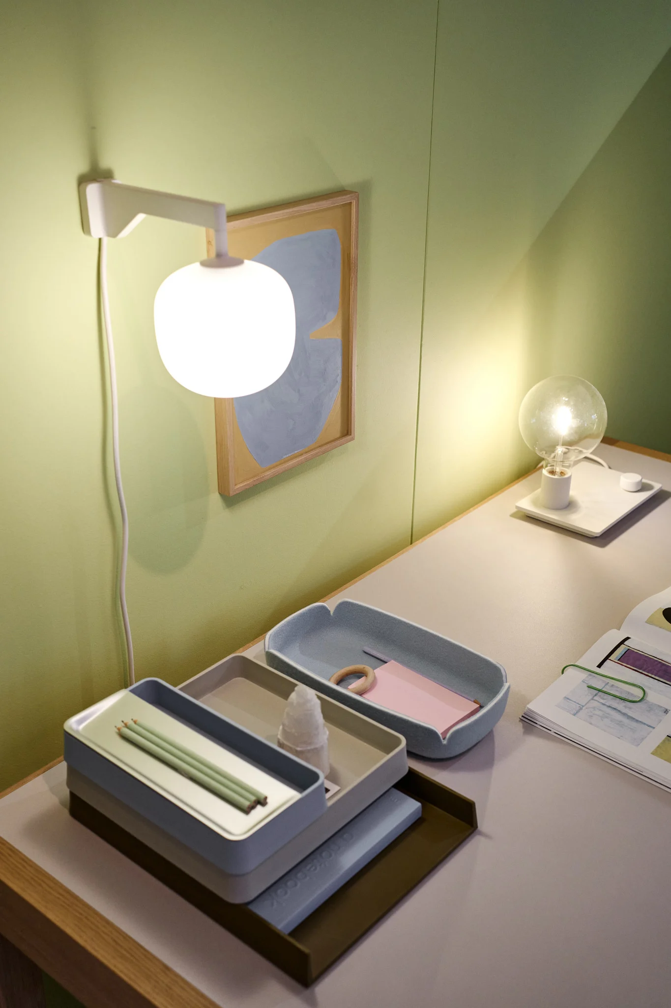 Ein ordentlicher Schreibtisch vor einer grünen Wand mit der beigefarbenen Rime Wandleuchte von Muuto.