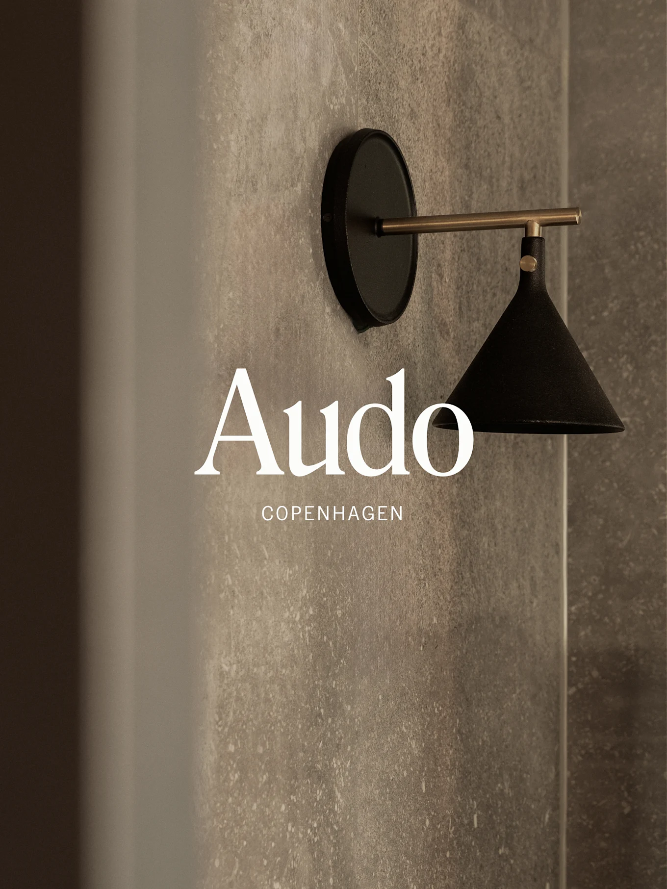 Eine wandmontierte schwarze Lampe mit einem Messingarm vor einer strukturierten grauen Wand, mit dem Text 'Audo Copenhagen'.