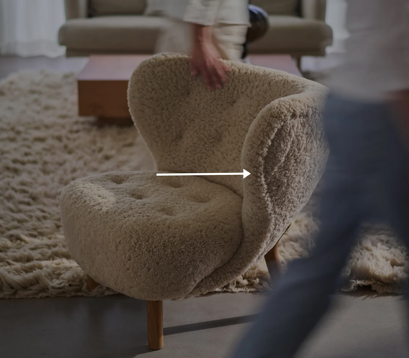 Cremefarbener, flauschiger Sessel mit Holzbeinen in einem Wohnzimmer, eine verschwommene Hand berührt die Rückenlehne.