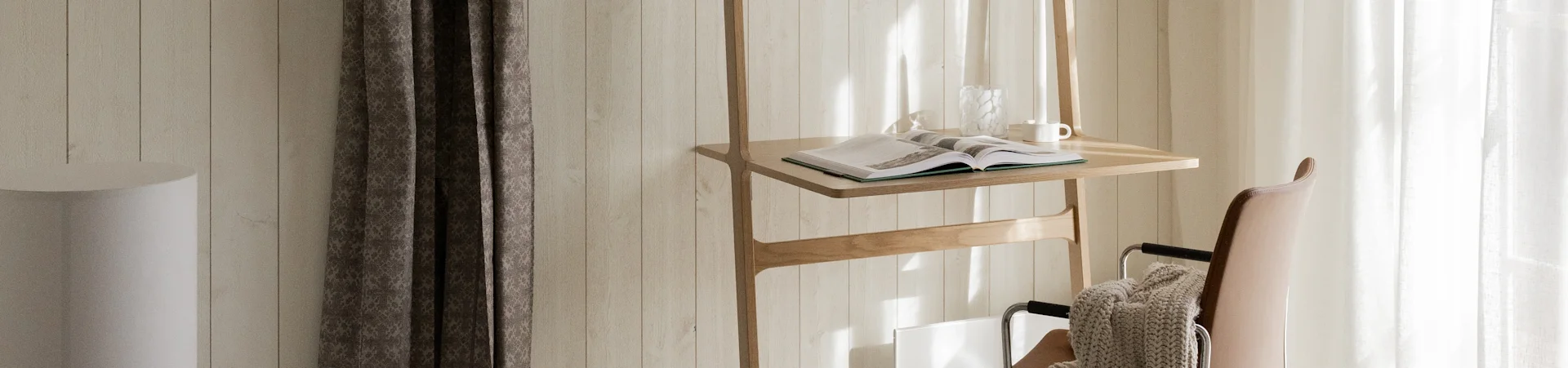 Ein Holzständer mit offenem Buch, Glasvase und weißem Becher, neben einem braunen Lederstuhl mit beiger Decke, im Sonnenlicht.