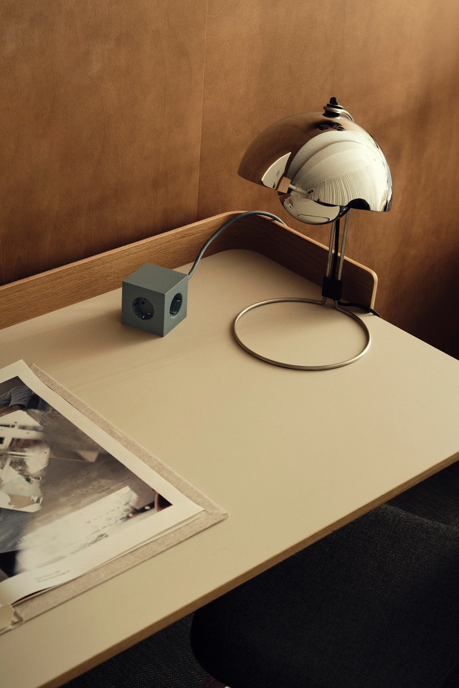 Ein minimalistisches Schreibtisch-Setup mit einer verchromten Kuppellampe, einer grauen Würfelsteckdosenleiste und einem offenen Buch auf einer hellen Oberfläche.