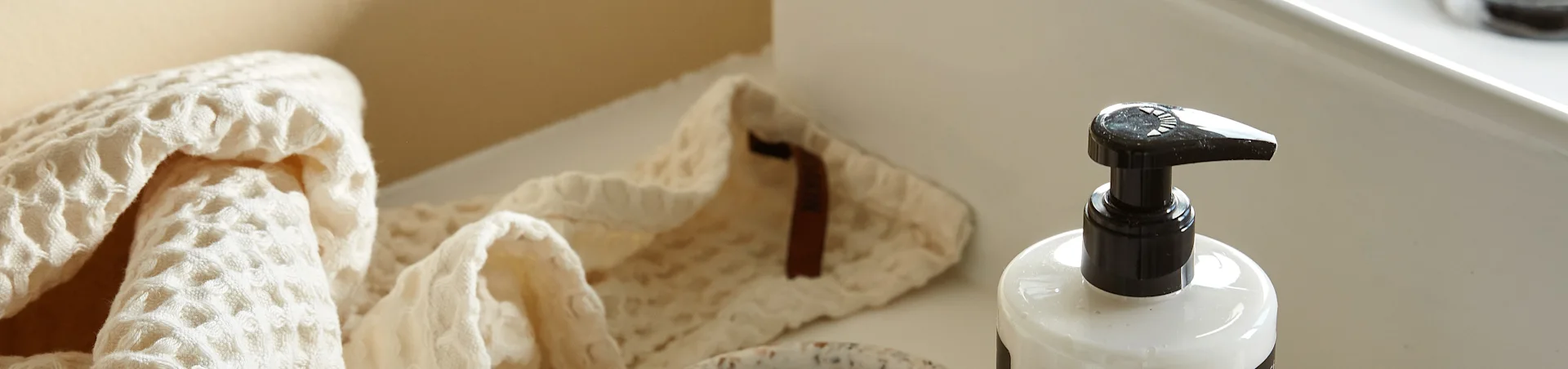 Ein helles, waffelgewebtes Handtuch, gefaltet neben einer weißen Pumpflasche auf einer sauberen, hellen Oberfläche.