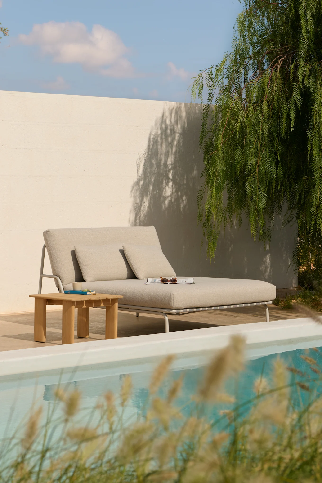 Hellgraue Outdoor-Chaiselongue mit Kissen, einem Holztisch und einer Zeitschrift am Pool.