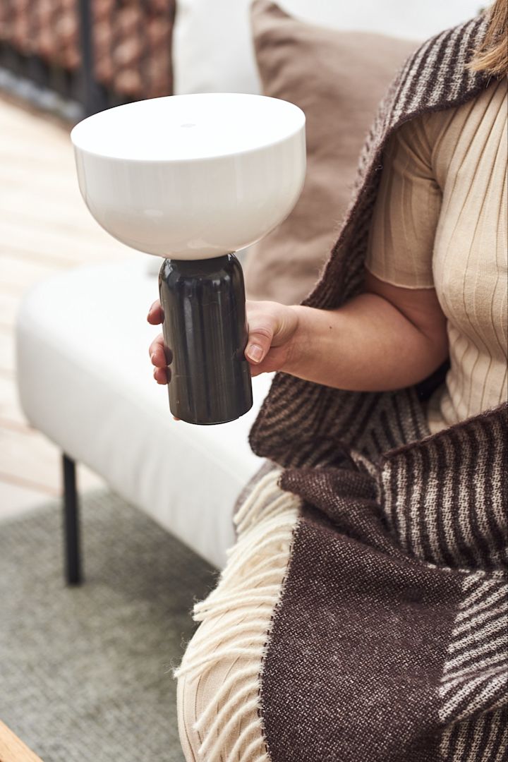 Die kabellose Kizu Tischleuchte wird von New Works wird von einer Person in der Hand gehalten, welche in eine Decke gewickelt draußen auf einem Outdoor-Sofa sitzt.