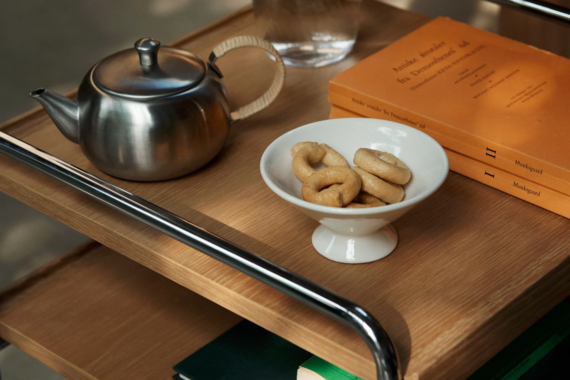 Eine Teekanne aus Edelstahl, eine weiße Schale mit Ringkeksen und zwei orangefarbene Bücher auf einem Holzregal.