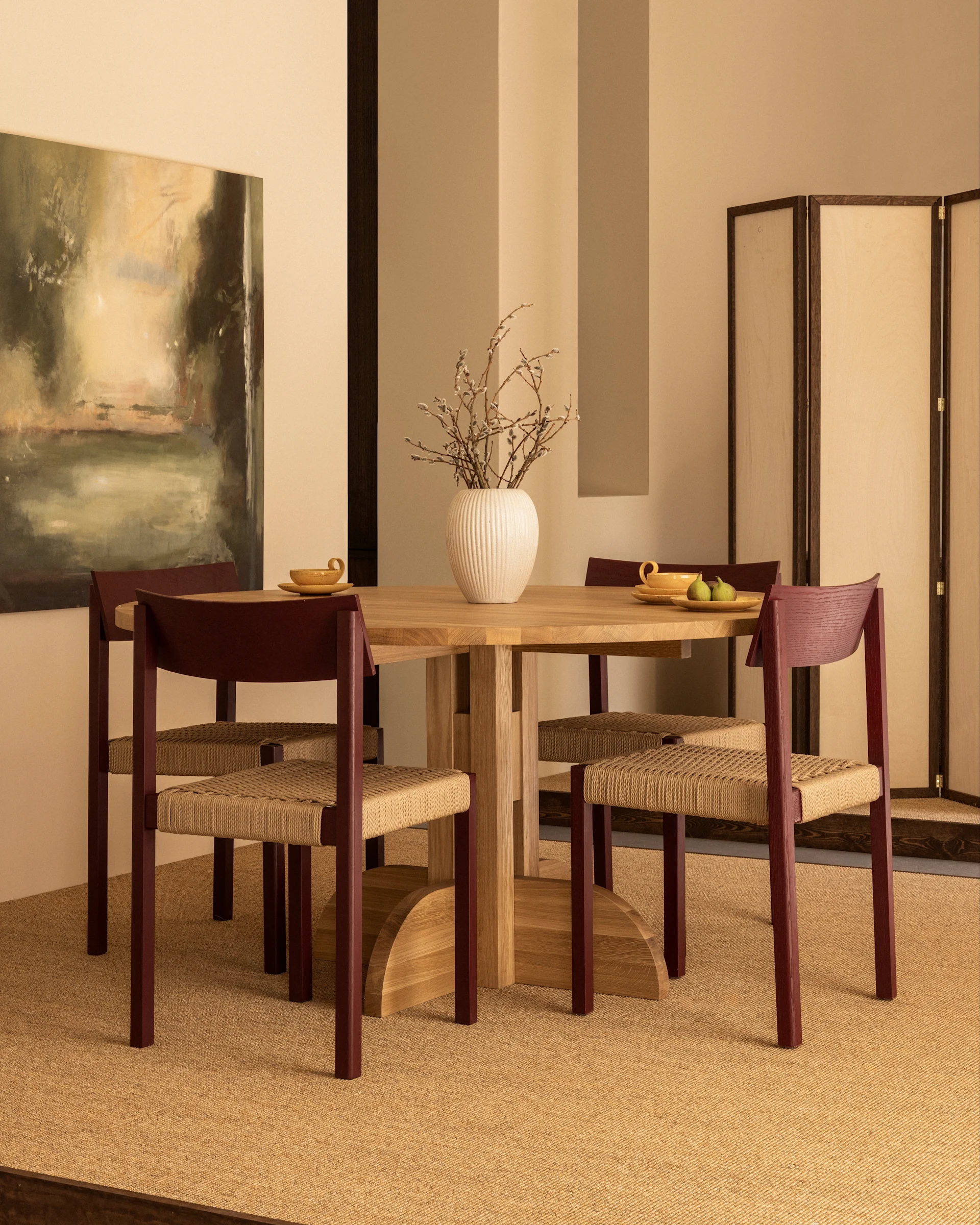Ein Esszimmer mit einem runden Holztisch, vier Stühlen mit rotbraunen Rahmen und geflochtenen Sitzen sowie einer Vase mit Weidenkätzchen.