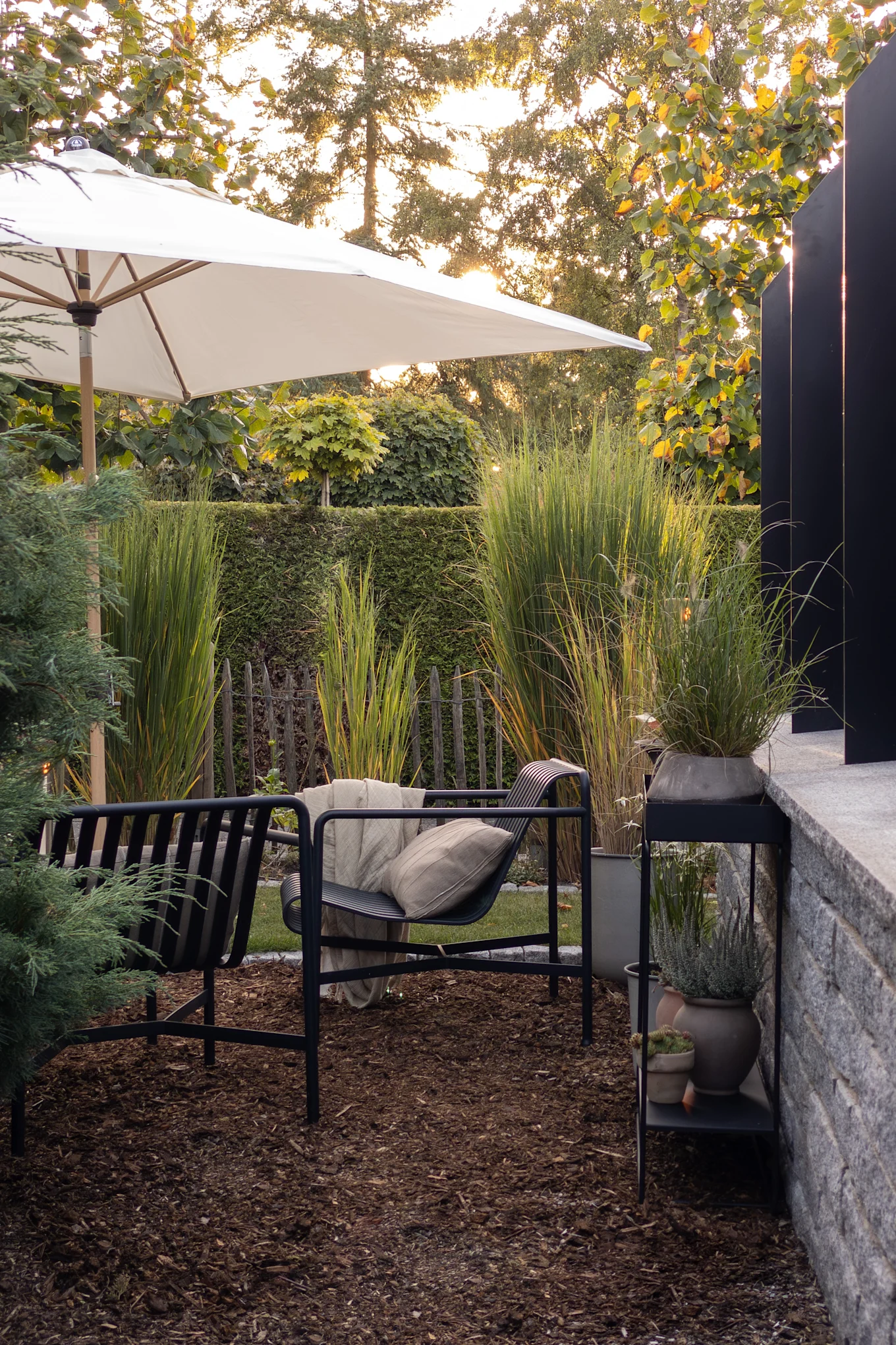 Außenbereich mit schwarzem Loungesessel, beigem Kissen und Decke unter einem weißen Sonnenschirm, umgeben von hohen Gräsern und Gartenpflanzen.