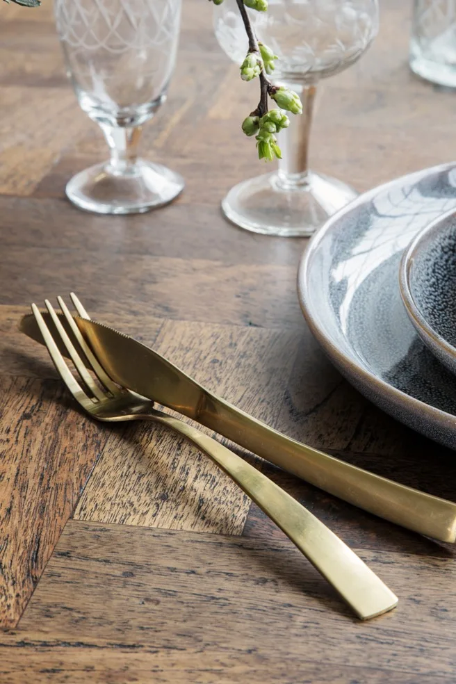 Gedeckter Tisch mit gestapelten grauen Keramiktellern, goldenem Besteck, klaren Ätzgläsern und einem Ast mit grünen Knospen.