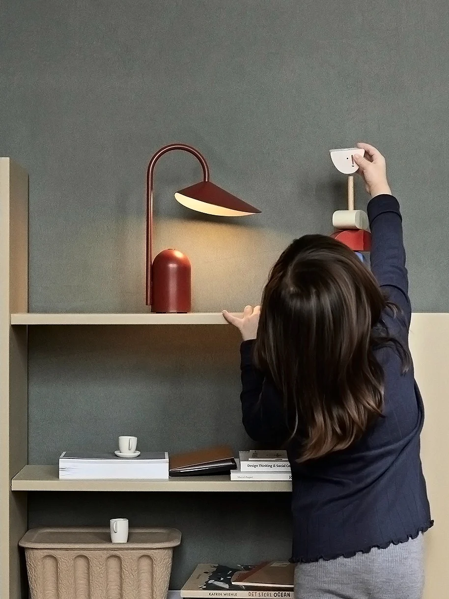 Ein Kind greift nach einem Holzspielzeugvogel auf einem hellen Regal mit einer roten Lampe und Büchern; ein geflochtener Korb steht auf dem Boden.