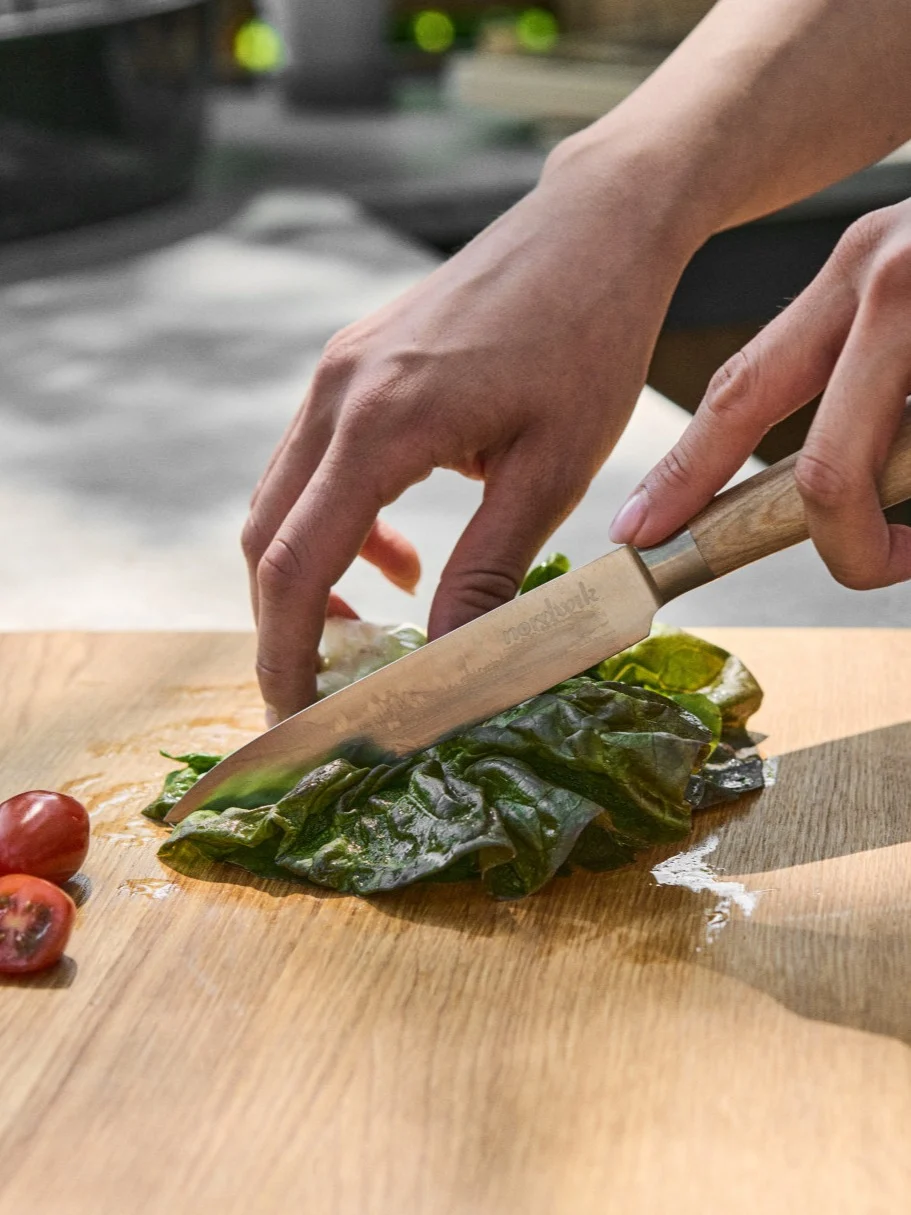 Hände schneiden frischen grünen Salat auf einem Holzschneidebrett mit einem Messer, daneben geschnittene Kirschtomaten.