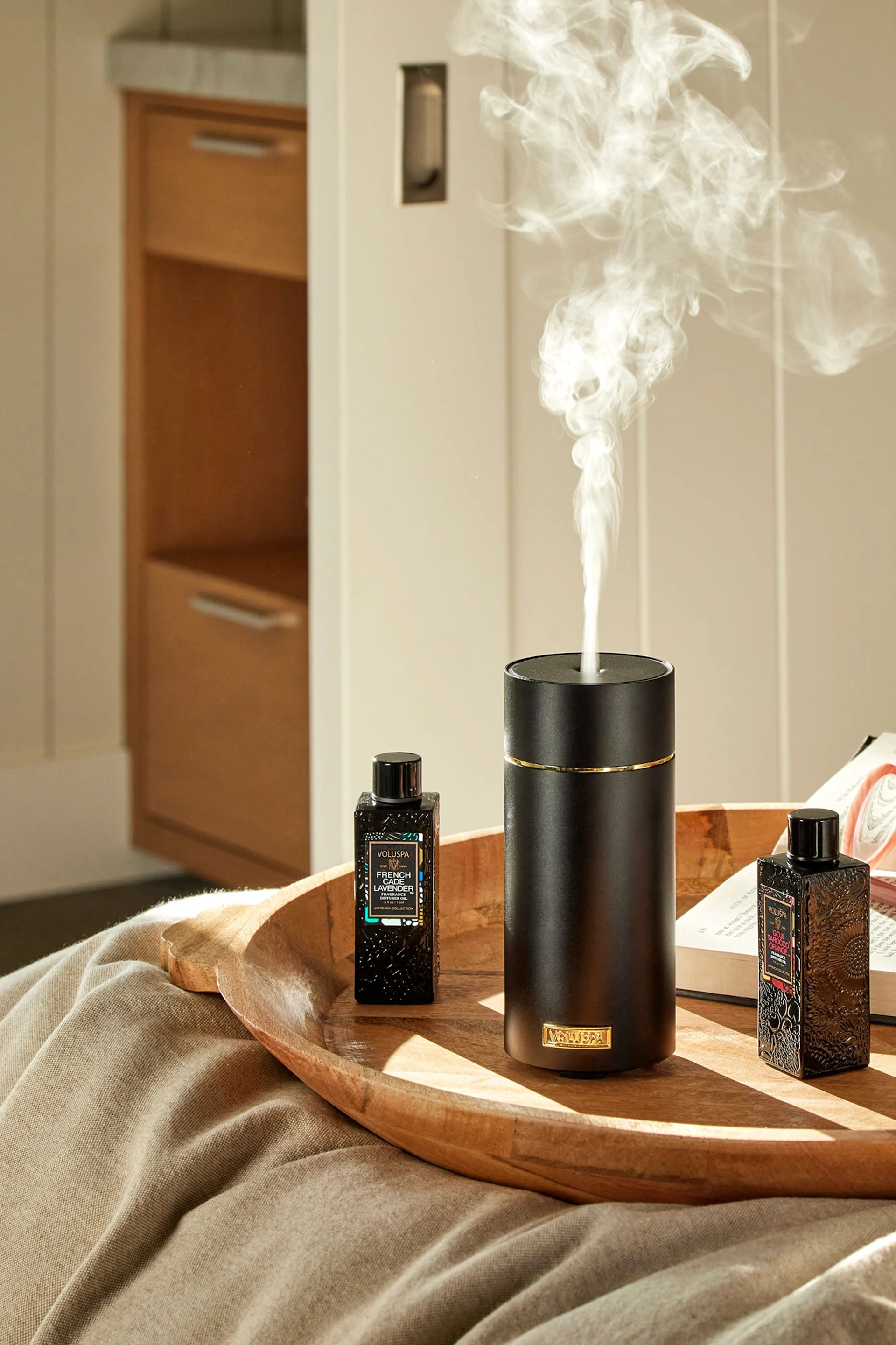 Der Japonica Ultrasonic Diffuser von Voluspa steht auf einem Tablett auf einem Bett, umgeben von verschiedenen Düften.