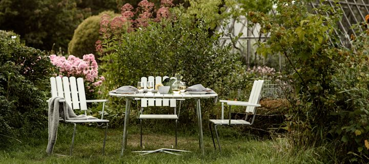 Grythyttan: Die Klassiker-Serie, bestehend aus drei Stühlen und einem Tisch, steht in einem gemütlichen Garten mit zahlreichen blühenden Blumen, im Hintergrund ein großes Gewächshaus.