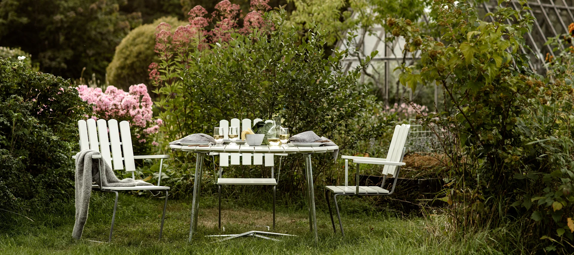 Grythyttan: Die Klassiker-Serie, bestehend aus drei Stühlen und einem Tisch, steht in einem gemütlichen Garten mit zahlreichen blühenden Blumen, im Hintergrund ein großes Gewächshaus.
