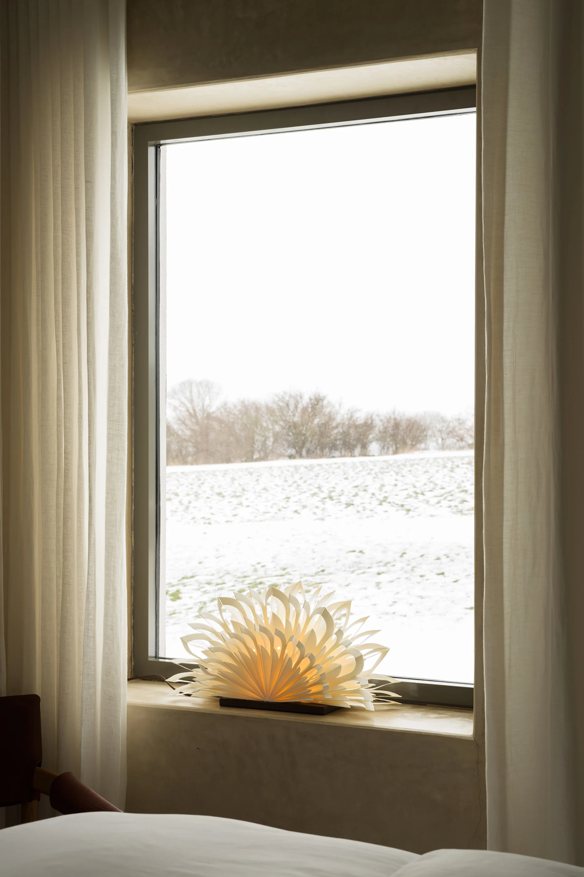 Fenster mit Blick auf ein verschneites Feld, weißen Vorhängen und einer leuchtenden Papierlampe auf dem Sims.