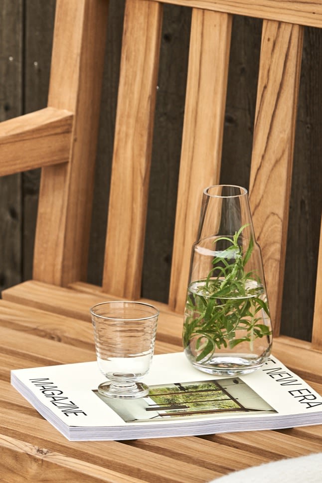 Die Viva Karaffe, gefüllt mit Wasser und Rosmarin, steht neben einem dazupassenden Glas auf einer Holzbank.