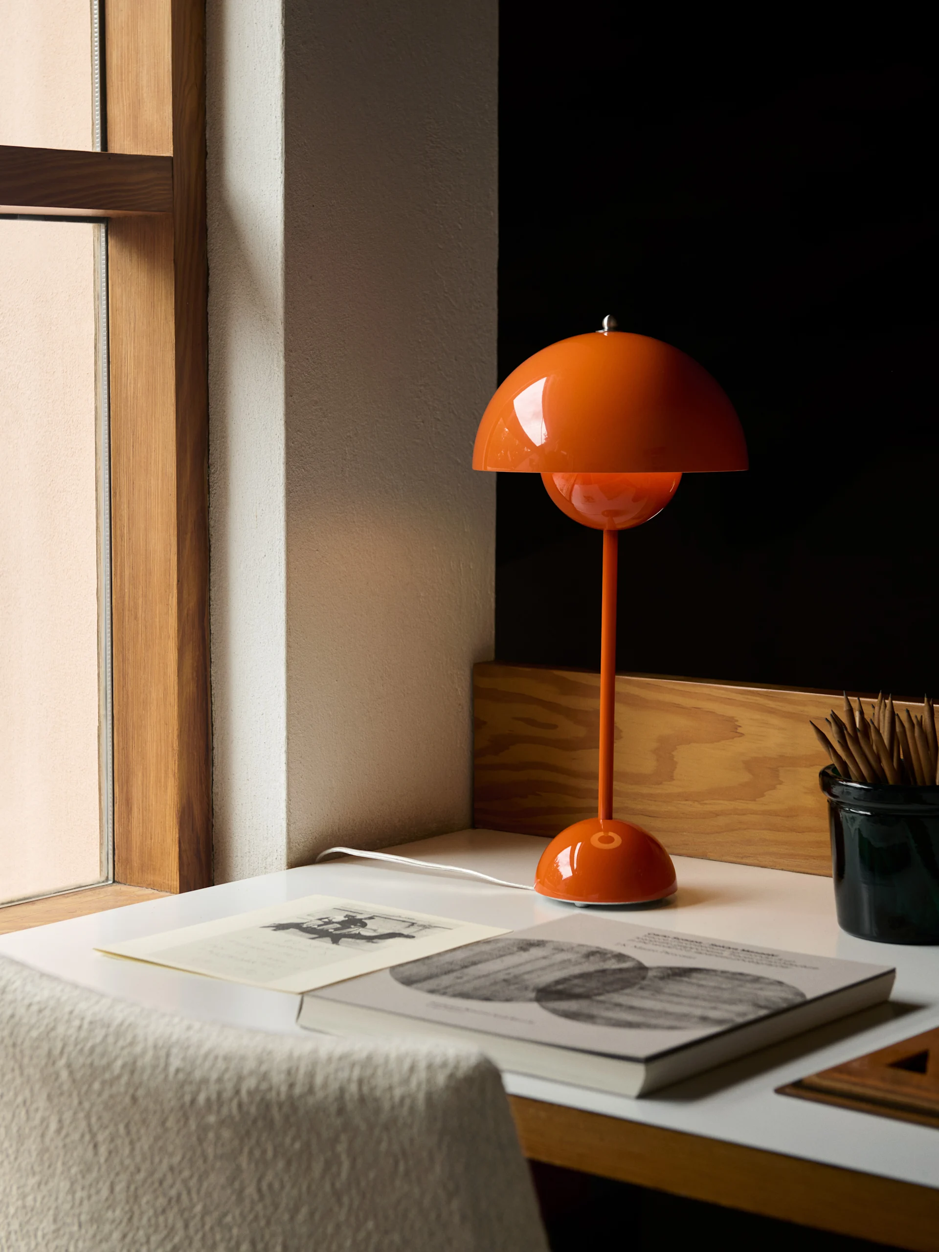 Eine orangefarbene, kuppelförmige Tischlampe steht auf einem weißen Schreibtisch neben Büchern und einem Bleistiftbehälter.