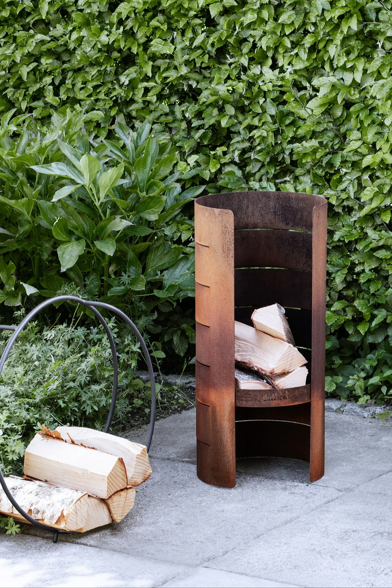 Ein rostiger Metall-Holzlager für draußen, gefüllt mit Brennholz, neben einem runden Holzhalter auf einer Terrasse vor einer grünen Hecke.