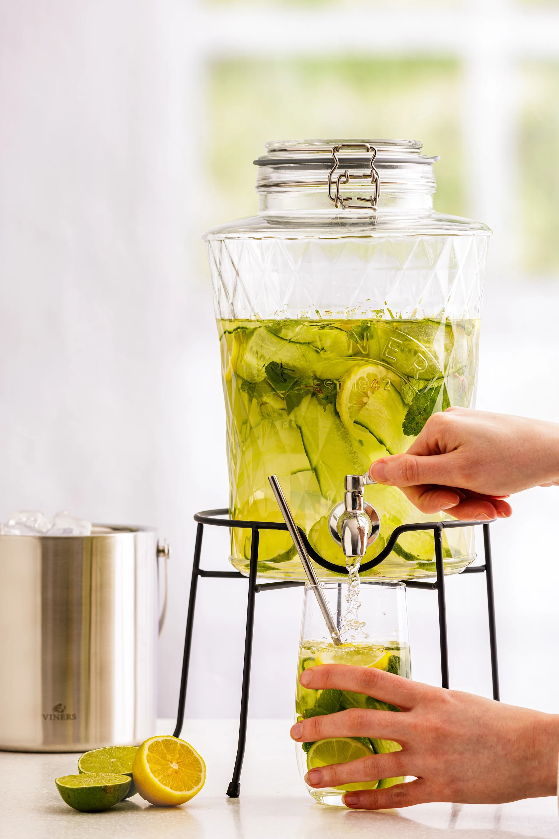Hände einer Person füllen ein Glas mit erfrischendem Gurken-, Zitronen- und Minzwasser aus einem klaren Glasbehälter. Ein Viners Eiswürfelbehälter und Zitrushälften sind in der Nähe.
