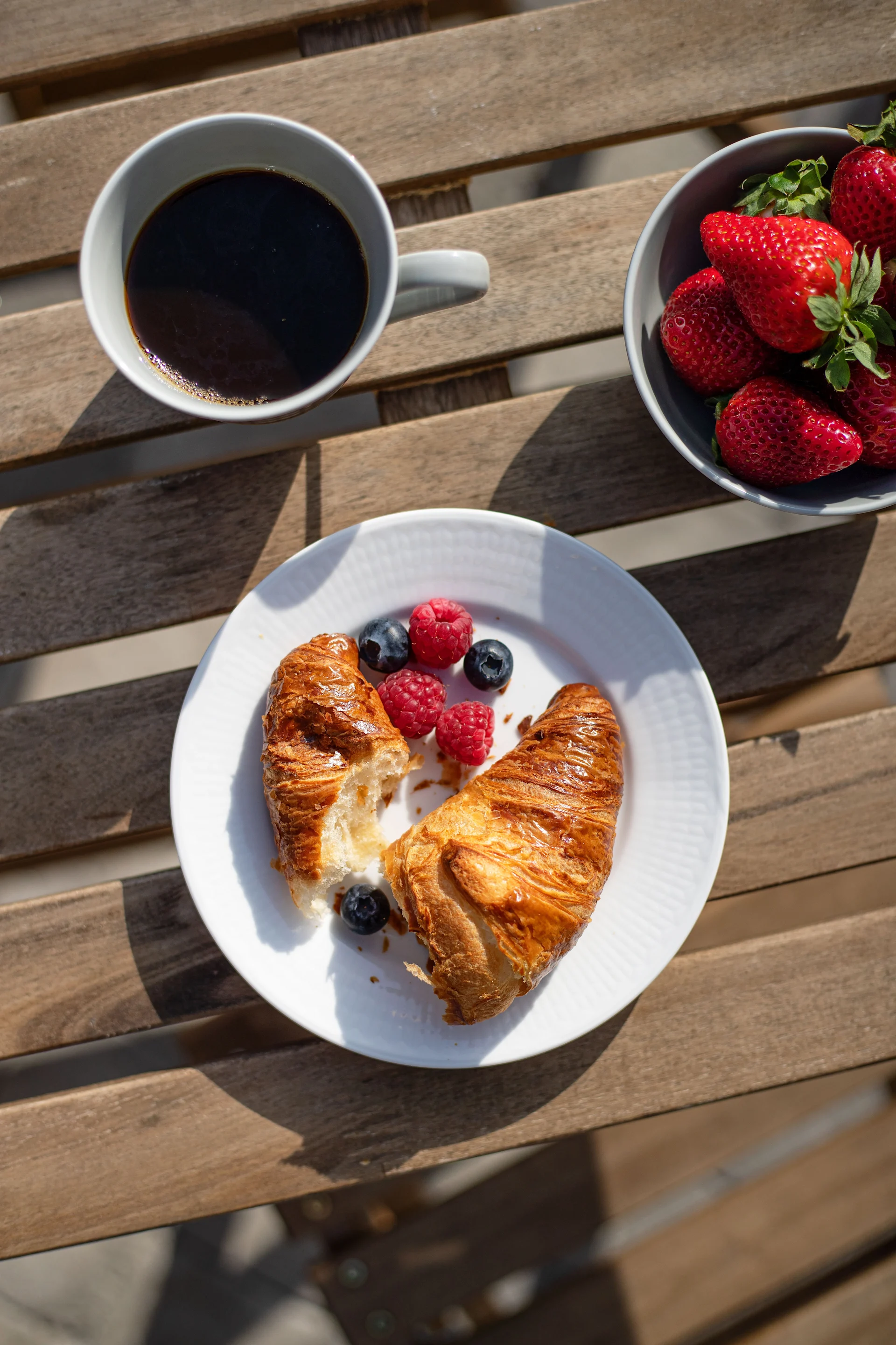 Draufsicht auf ein Frühstück auf einem Holztisch mit Kaffee, Erdbeeren, Croissants, Heidelbeeren und Himbeeren.