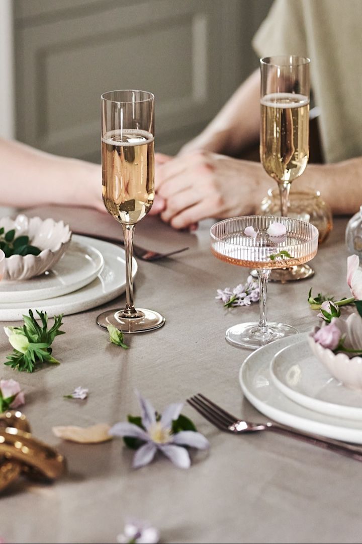 Zu Ihrem romantischen Essen zu Hause können Sie den Tisch auch verspielt dekorieren. Hier sehen Sie weißes Geschirr, schlichte Gläser und Blüten auf einer beigefarbenen Tischdecke.