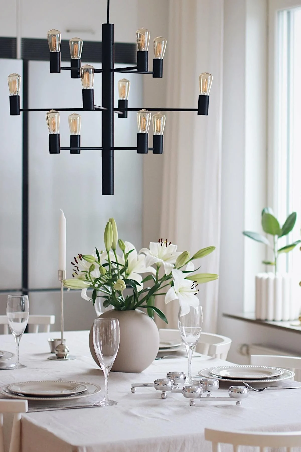 11 stylische Pendelleuchten für Ihr Wohlfühlzuhause - hier sehen Sie den Manola 12 Kronleuchter von Herstal über einem Strauß frischer Blumen in einem beigen Zimmer hängen.