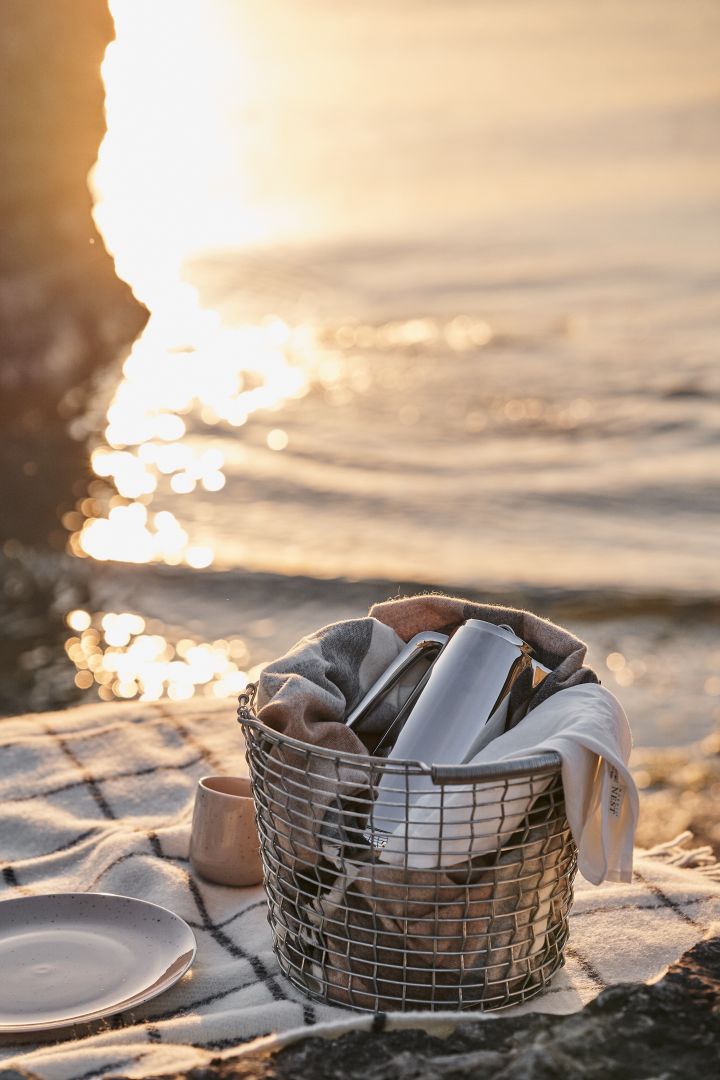 Ein Must Have ist der robuste Korbo-Metallkorb, der überallhin mitgenommen werden kann, hier am Strand gefüllt mit einer Thermoskanne und einer gemütlichen Decke.
