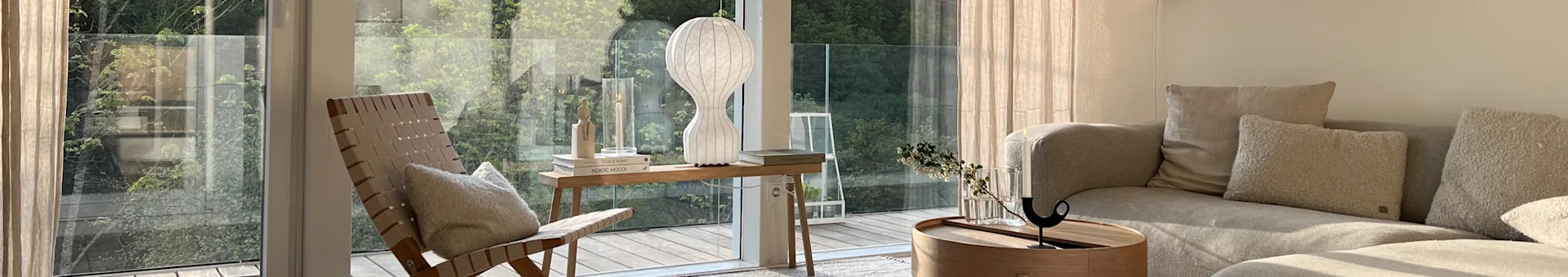 Sonnenhelles Wohnzimmer: Fenster, grüne Bäume. Geflochtener Stuhl, Konsole, hohe weiße Lampe, beiges Sofa, Couchtisch mit Blumen.