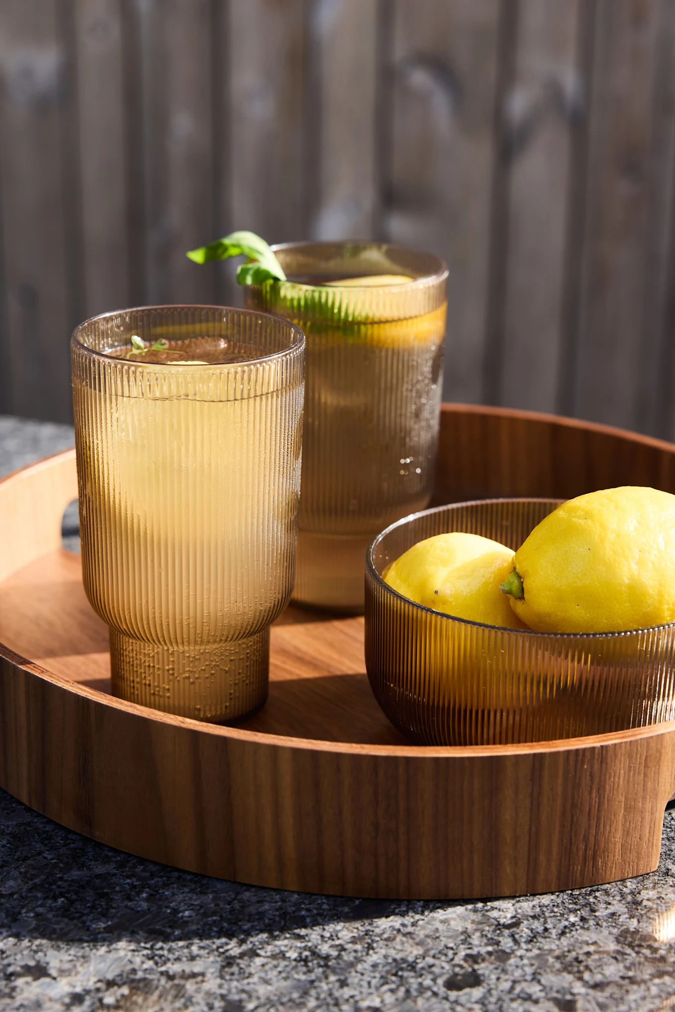 Zwei gerippte goldbraune Gläser mit Limonade und eine Schale Zitronen auf einem Holztablett im Freien.