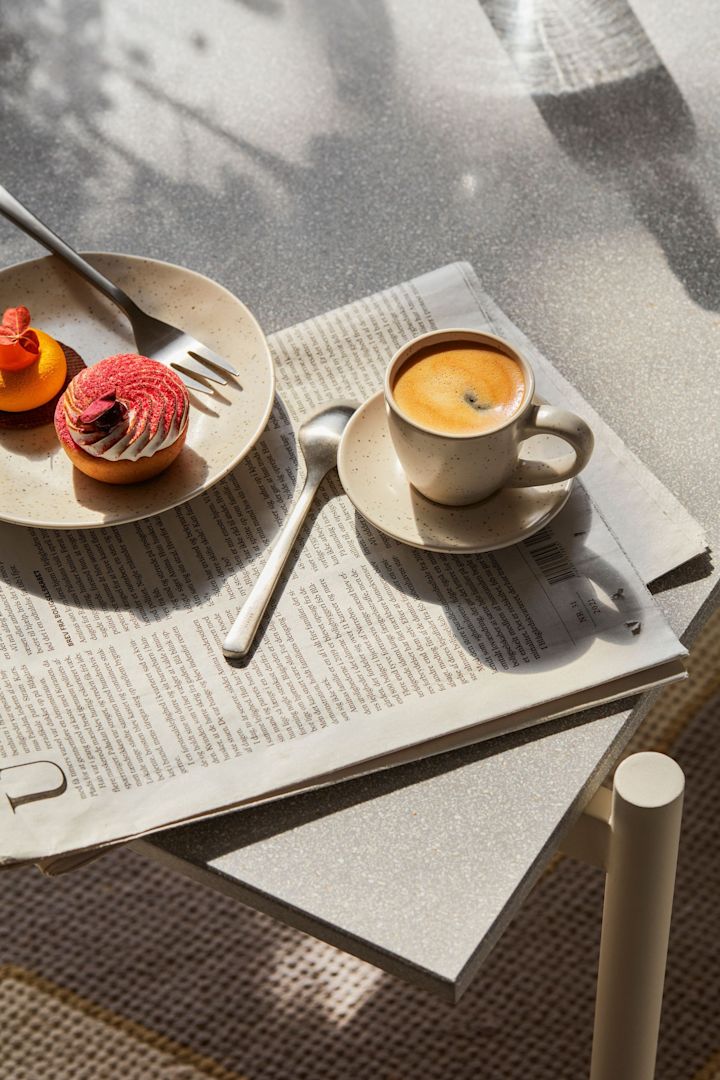Die Nordic Vanilla Espressotasse von Broste Copenhagen auf einem Frühstückstisch, darunter eine Tageszeitung, daneben ein Teller mit Kuchen.