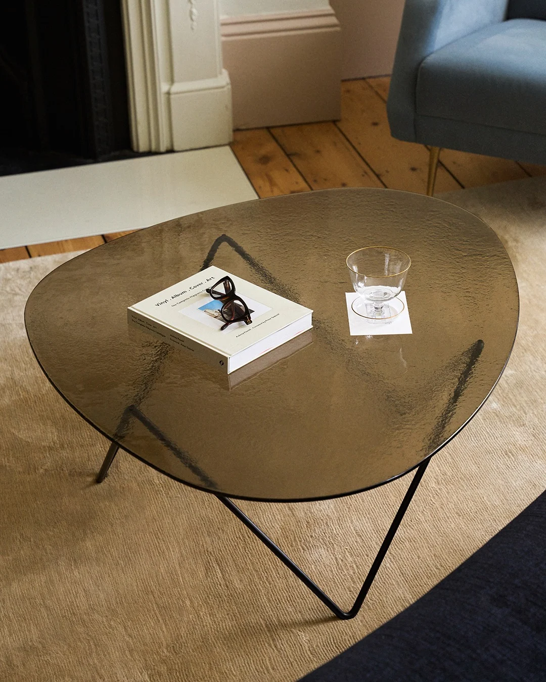 Organisch geformter Couchtisch aus dunklem Glas mit einem Buch, Schildpatt-Brille und einem Glas mit Goldrand auf einem Untersetzer.