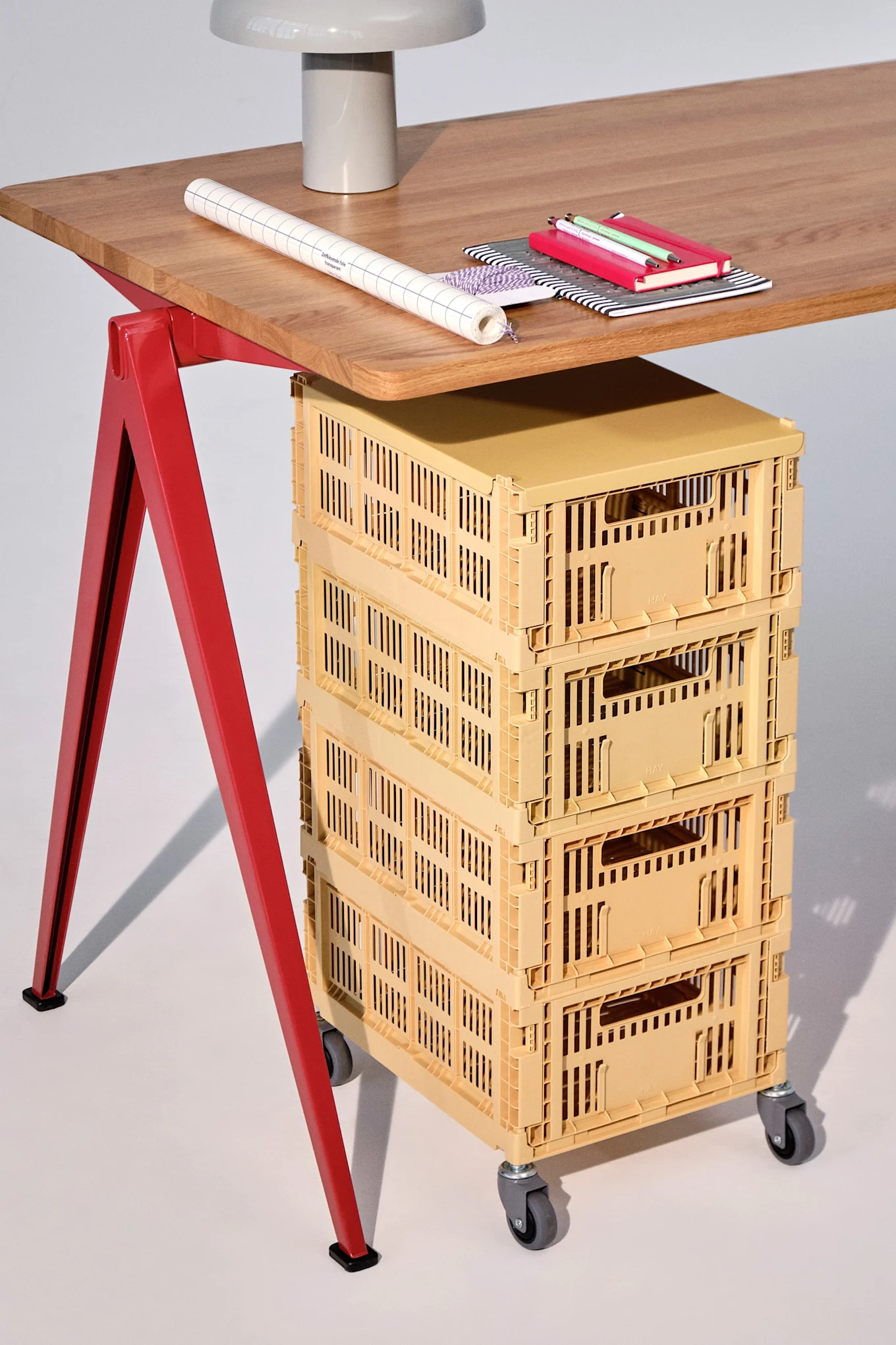 Holzschreibtisch mit roten Beinen, Lampe, Papier, Notizbüchern und Stiften, sowie vier gelben Rollcontainern darunter.