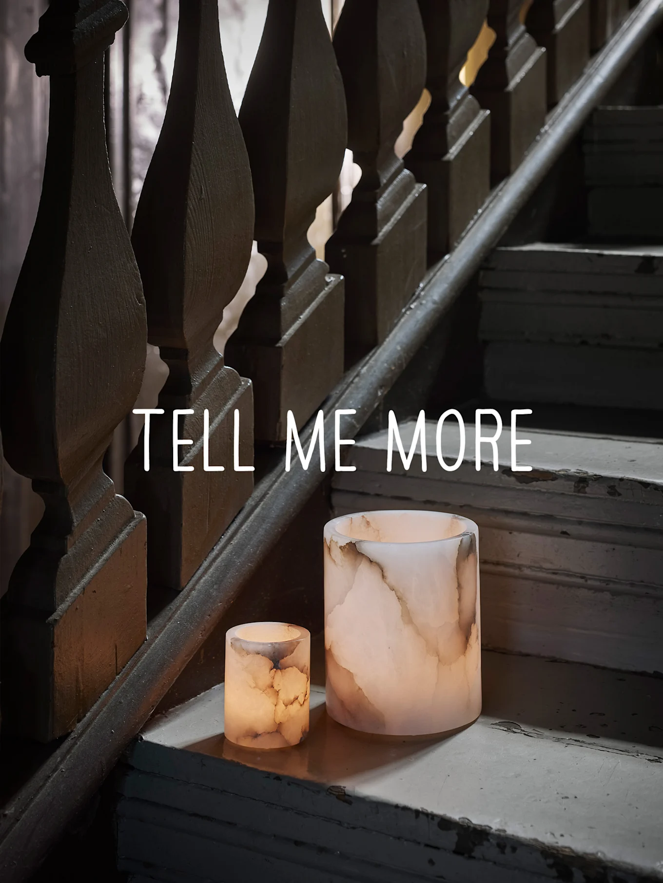 Zwei leuchtende onyxfarbene Kerzenhalter auf einer abgenutzten Holztreppe, mit dem Text "TELL ME MORE".