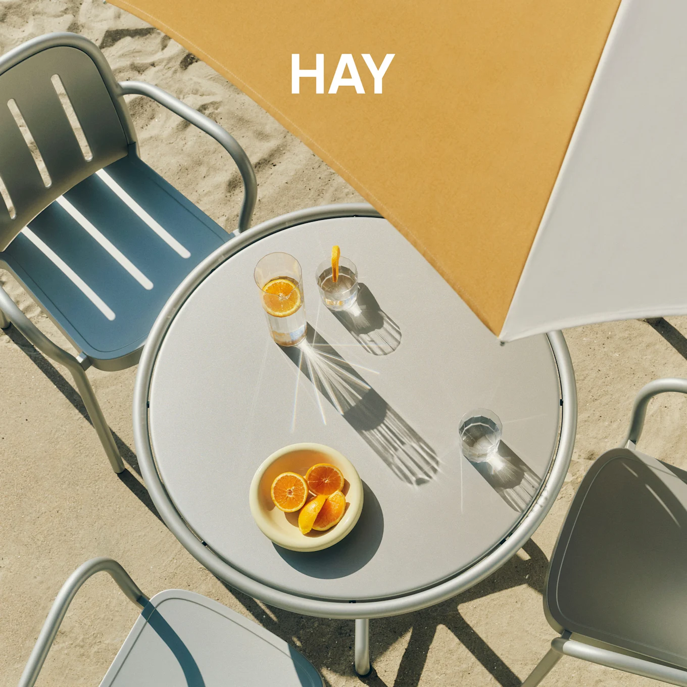 Aufsicht auf einen runden grauen Gartentisch mit zwei Stühlen auf Sand, darauf Getränke und Orangen unter einem gelb-weißen Sonnenschirm mit 'HAY'.