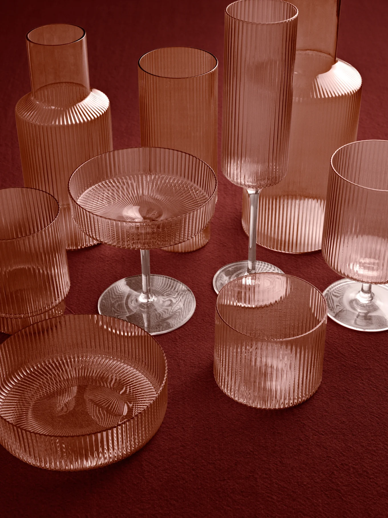 Sammlung gerippter, rötlich-brauner Glasvasen, Becher und Stielschalen auf einer tiefroten Oberfläche.