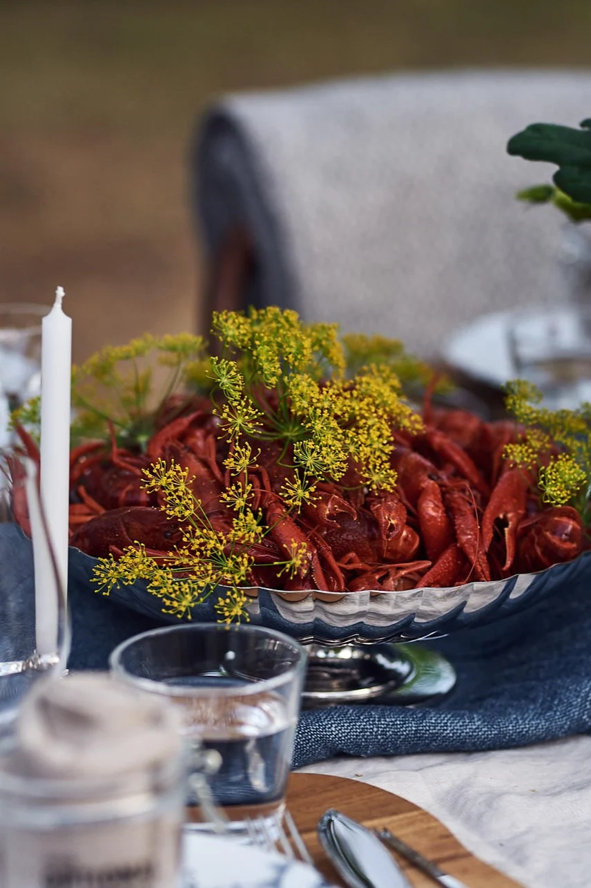 Im Freien gedeckter Tisch mit einer Silberschale voller roter Flusskrebse und Dill, Kerzen und einem Blumenarrangement.
