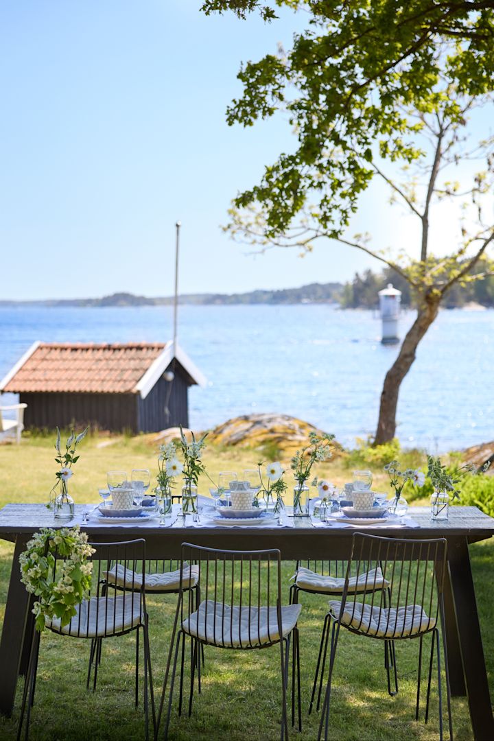 Ein mit Geschirr von Mateus in Blau-Weiß gedeckter Tisch im Grünen mit Aussicht auf ein kleines Haus und einen See.
