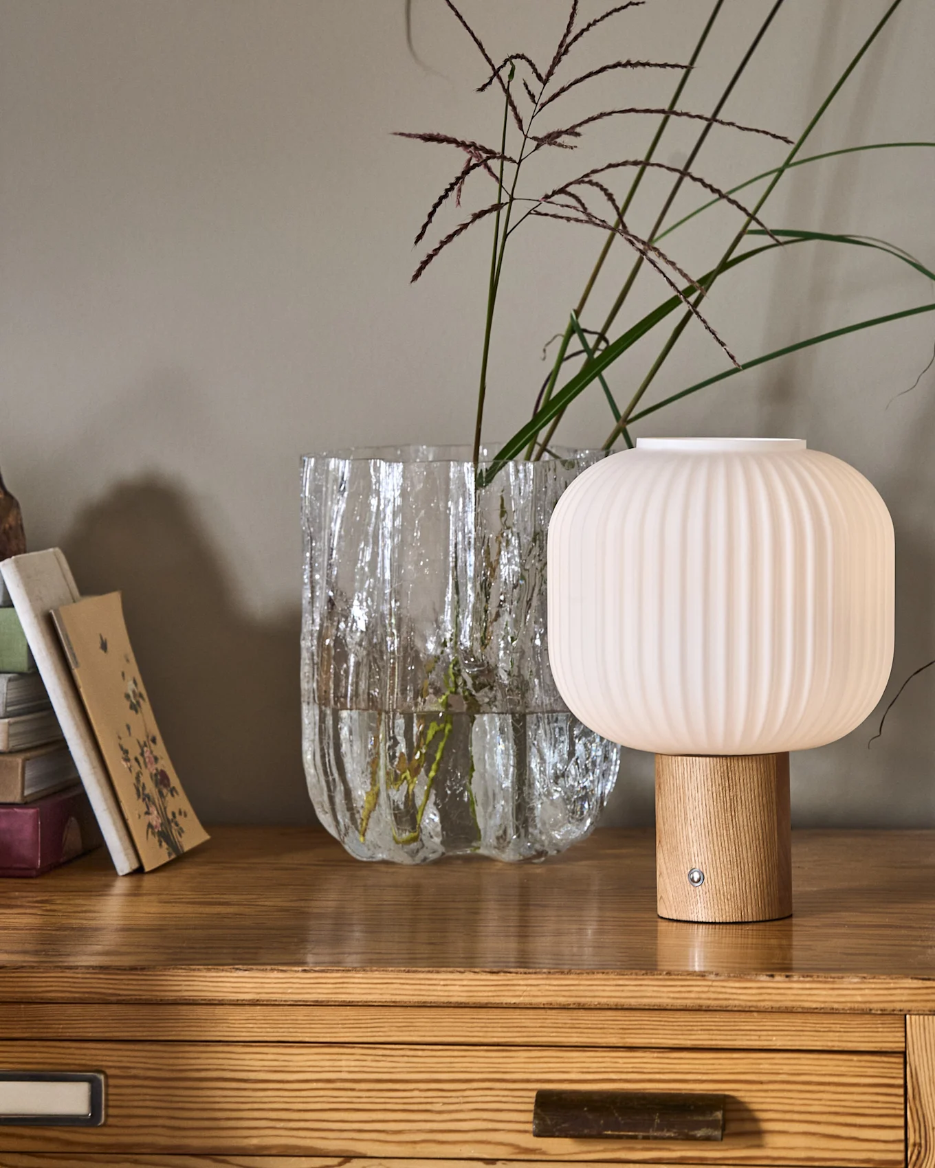 Eine geriffelte, pilzförmige Lampe mit Holzfuß neben einer strukturierten Glasvase mit Pflanzen und einem Bücherstapel auf einer Holzkommode.