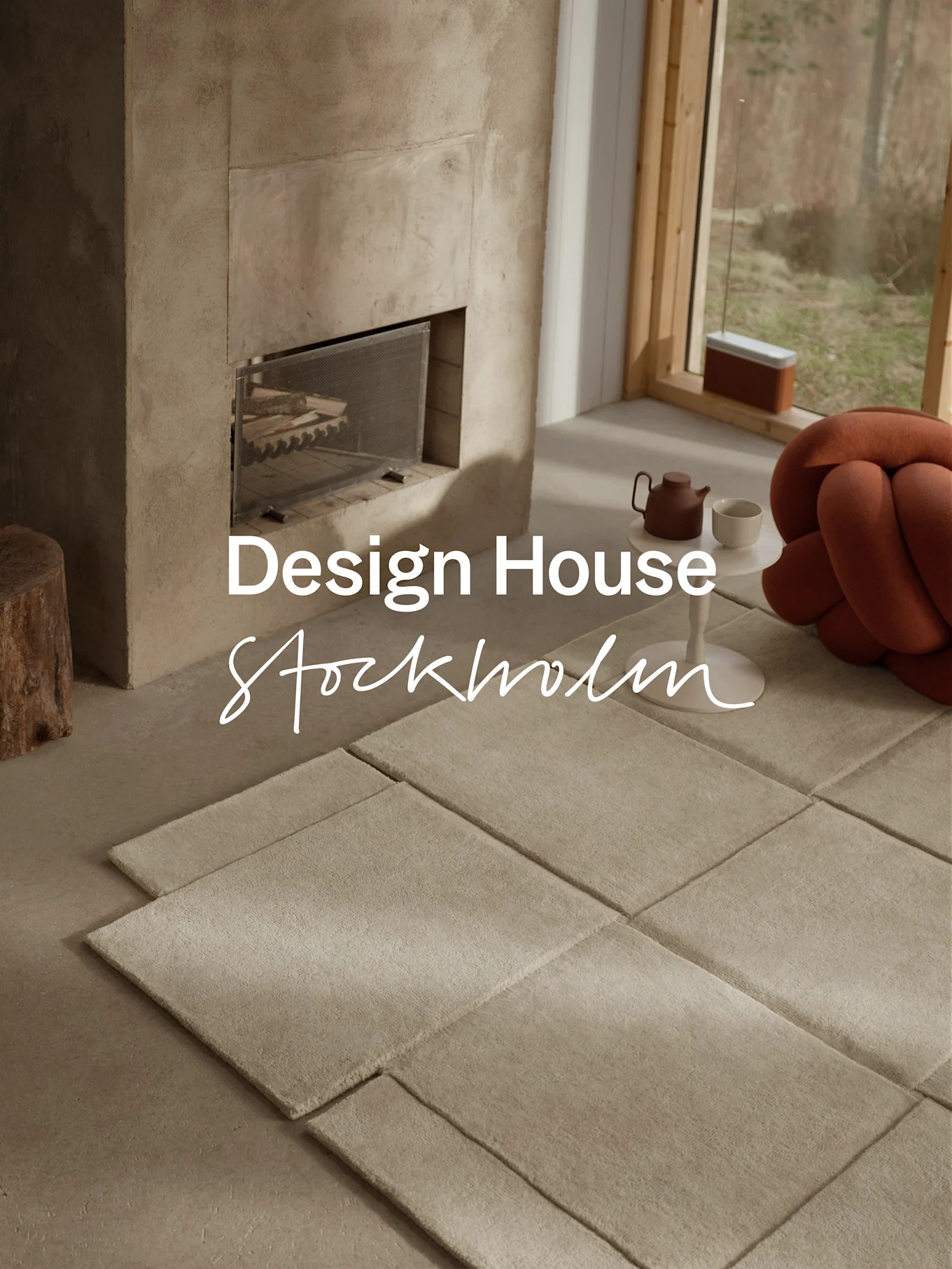 Modernes Wohnzimmer mit Beton-Kamin, modularen hellen Teppichen, einer Teekanne und einem orangefarbenen geknoteten Kissen neben einem großen Fenster.