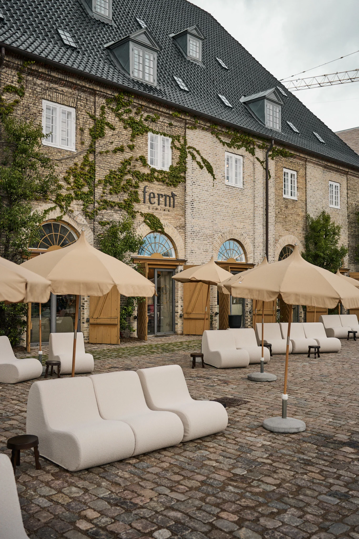 Outdoor-Sitzbereich mit cremefarbenen Modulsofas und beigen Schirmen auf Kopfsteinpflaster vor Backsteingebäude.