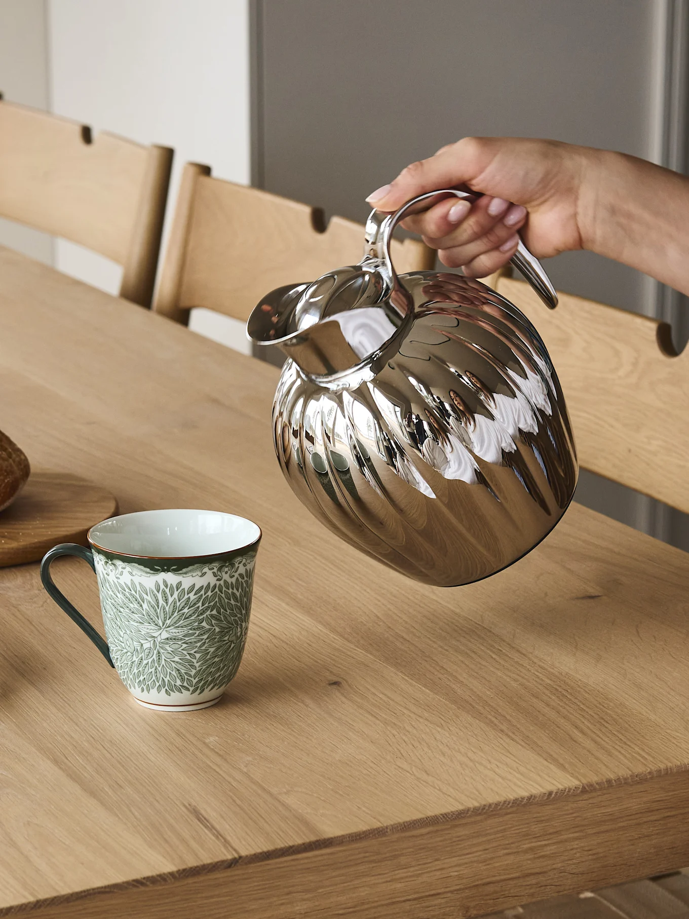 Eine Hand gießt aus einer silbernen geriffelten Thermoskanne in einen grün-weiß gemusterten Becher auf einem Holztisch.