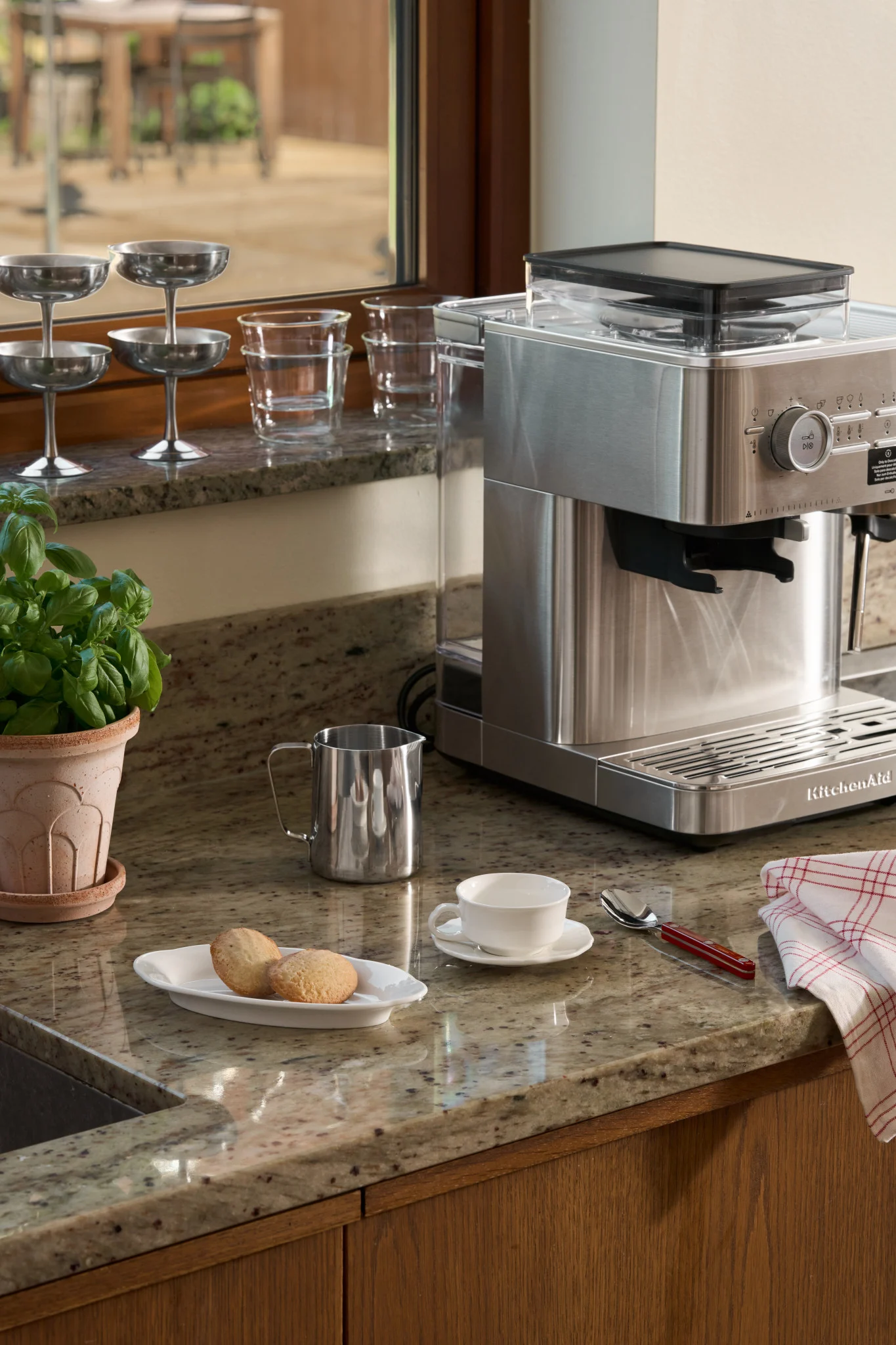 Eine Kaffeemaschine aus gebürstetem Edelstahl auf einer Granittheke mit einer weißen Tasse, Keksen, Basilikum und Milchkännchen.
