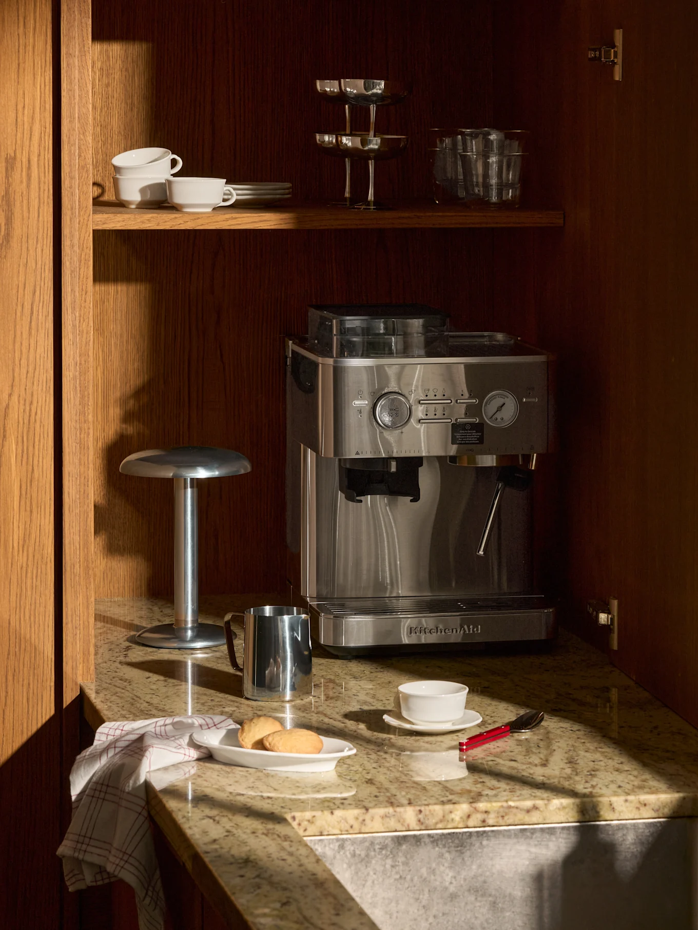 Eine Edelstahl-Espressomaschine auf Granitplatte im Holzschrank. Darüber Tassen und Gläser. Auf der Platte Kekse, Tasse und Milchkännchen.