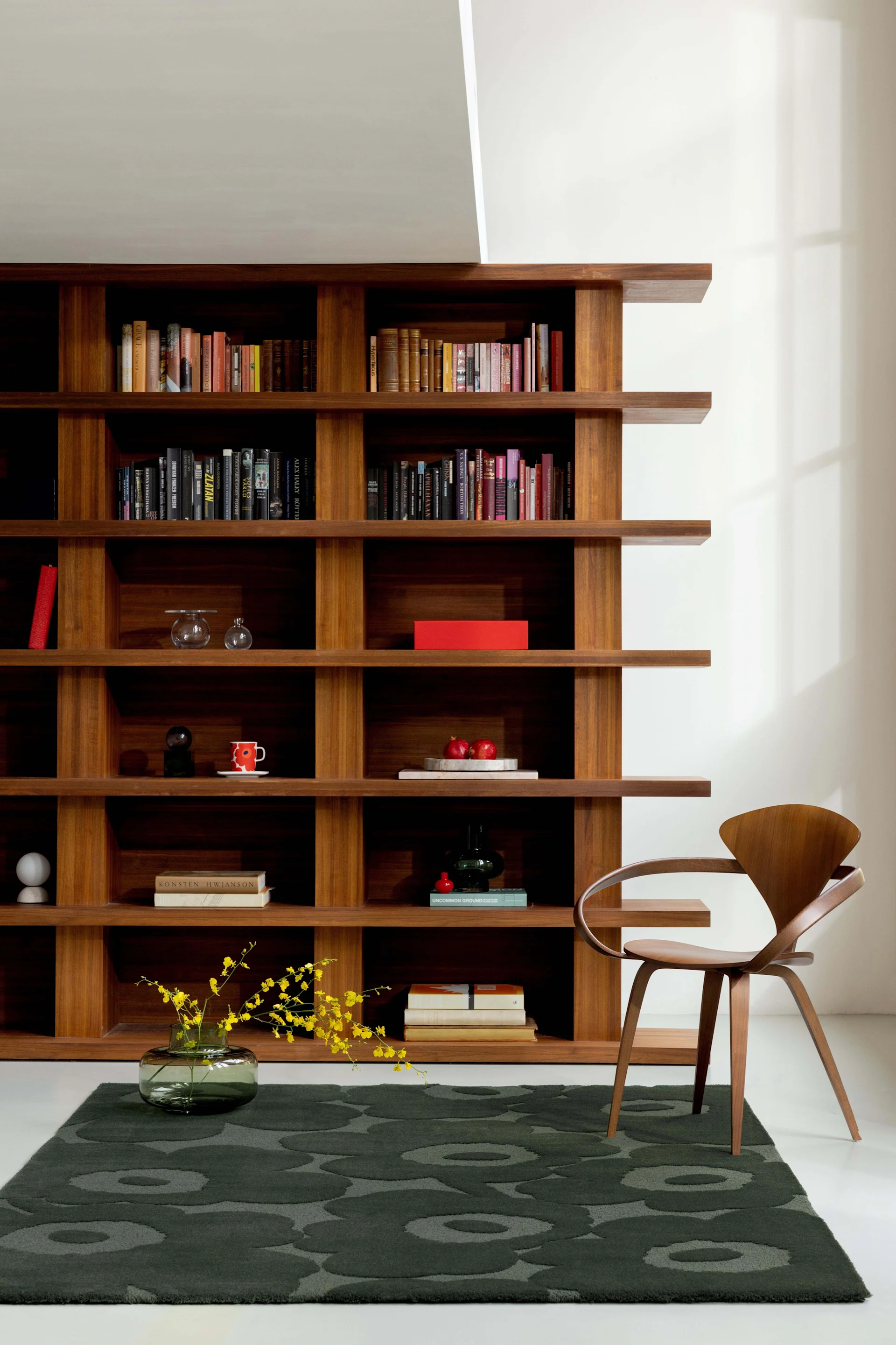 Ein stilvolles Zimmer mit einem hohen Holzregal voller Bücher und Dekoration, einem geschwungenen Holzstuhl und einem dunkelgrünen Teppich.