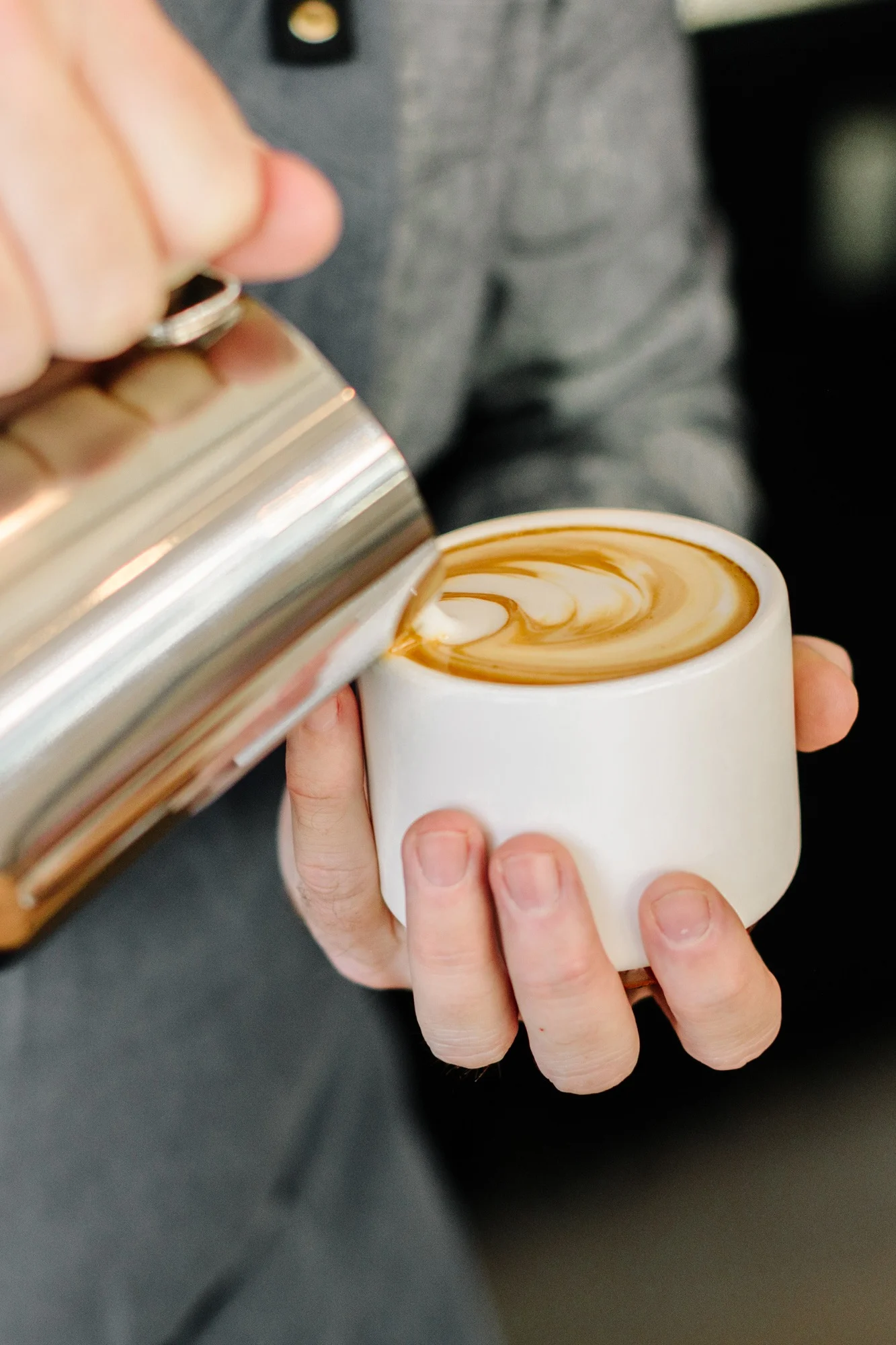 Nahaufnahme der Hände eines Barista, der Milch aus einem Milchkännchen in eine weiße Tasse gießt und Latte Art kreiert.